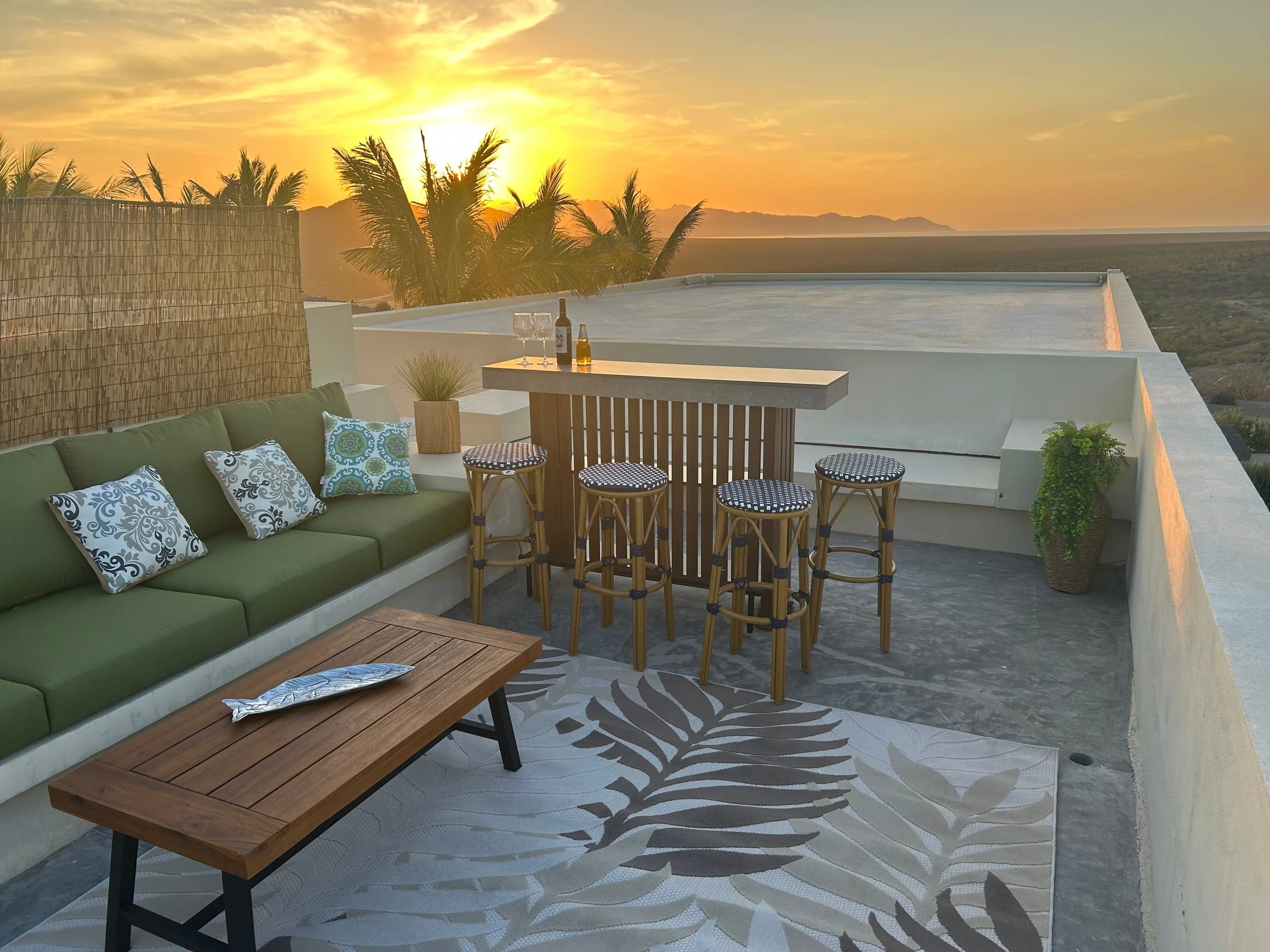 Rooftop patio with a green couch, decorative pillows, a wooden coffee table, a bar with four high stools, and a sunset view of palm trees and terrain.
