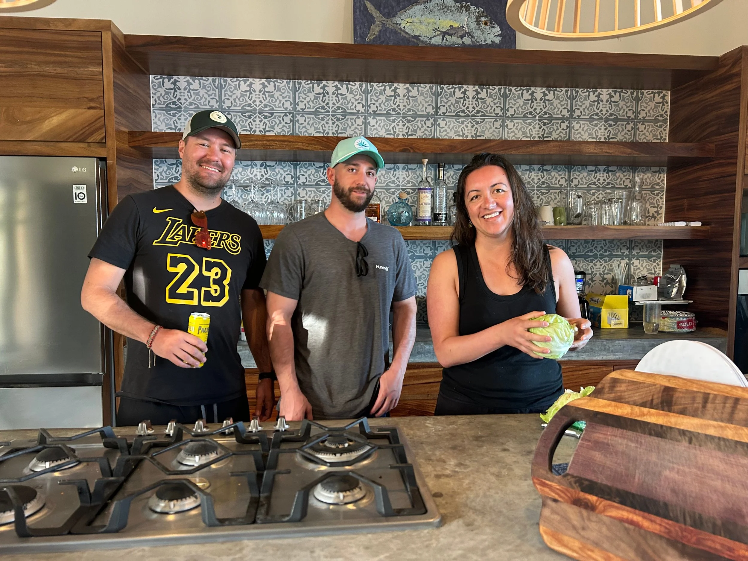 Three people standing in a kitchen smiling at the camera. The person on the left holding a can, the woman on the right holding a head of cabbage. The kitchen has a modern decor with open wooden shelves, a patterned backsplash, and a gas stove in the foreground.