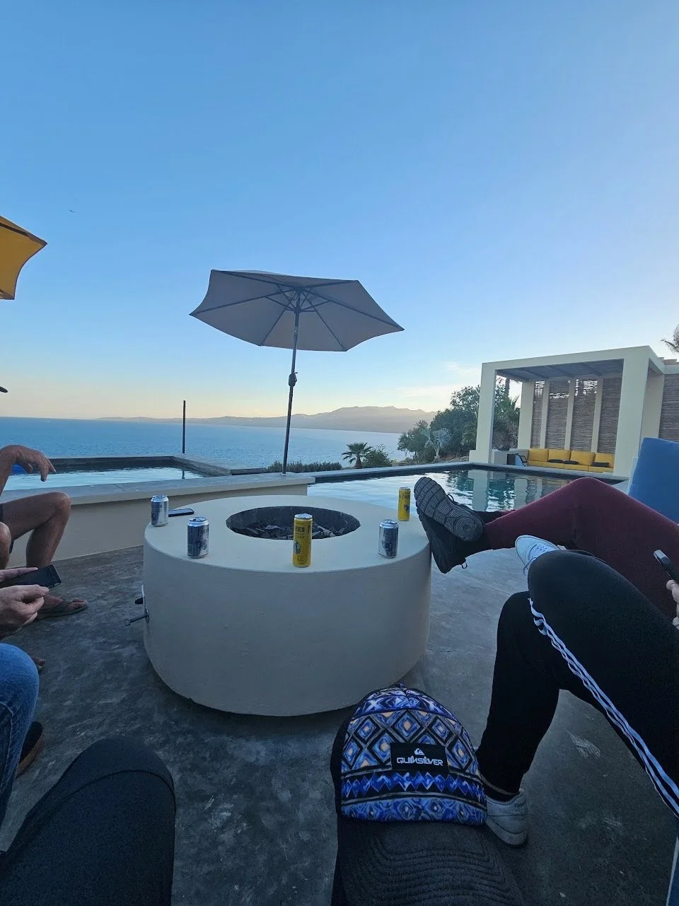 People sitting around a poolside with drinks, overlooking the ocean, with a large umbrella providing shade and modern outdoor furniture.