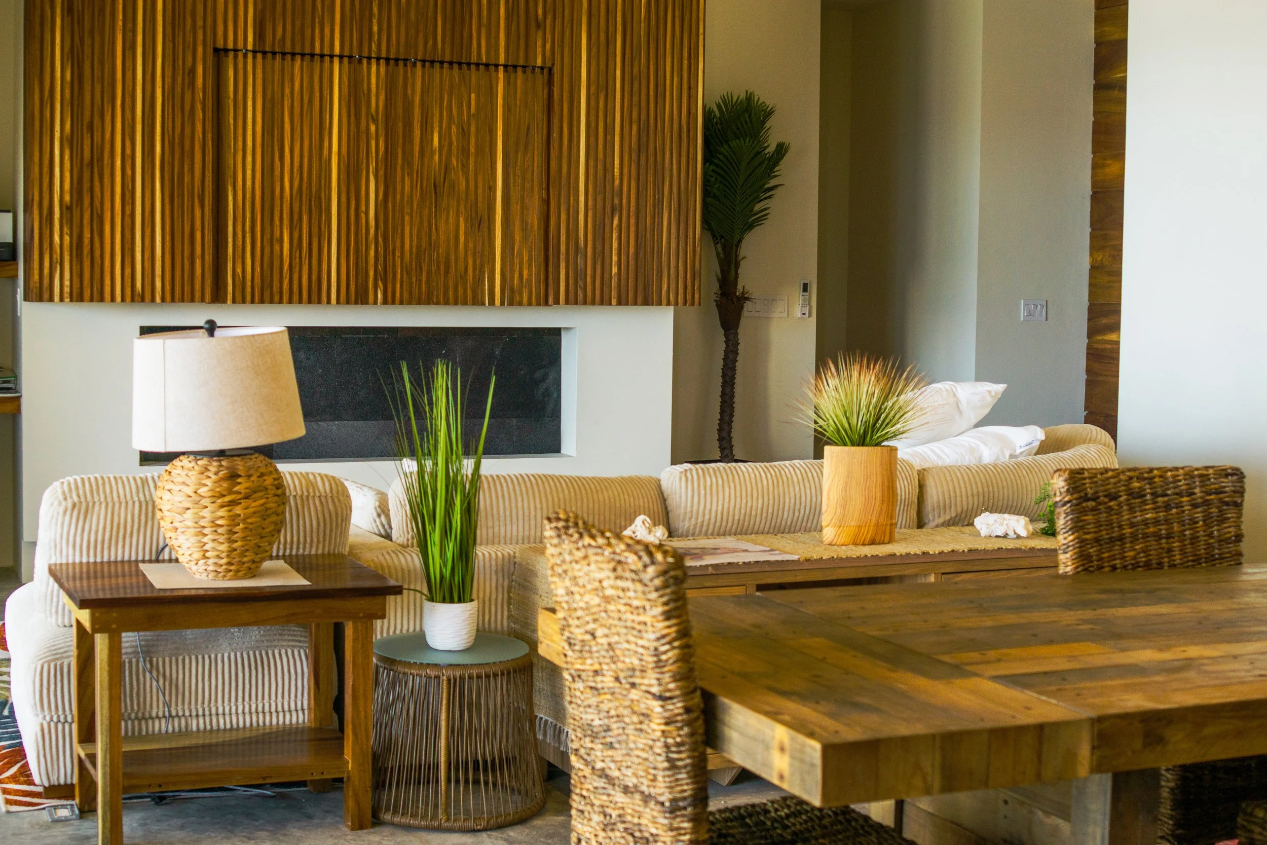 Living room with beige striped sofa, wooden side table with wicker lamp, tall plant, and decorative indoor plants on the table, with wooden paneling on the wall.