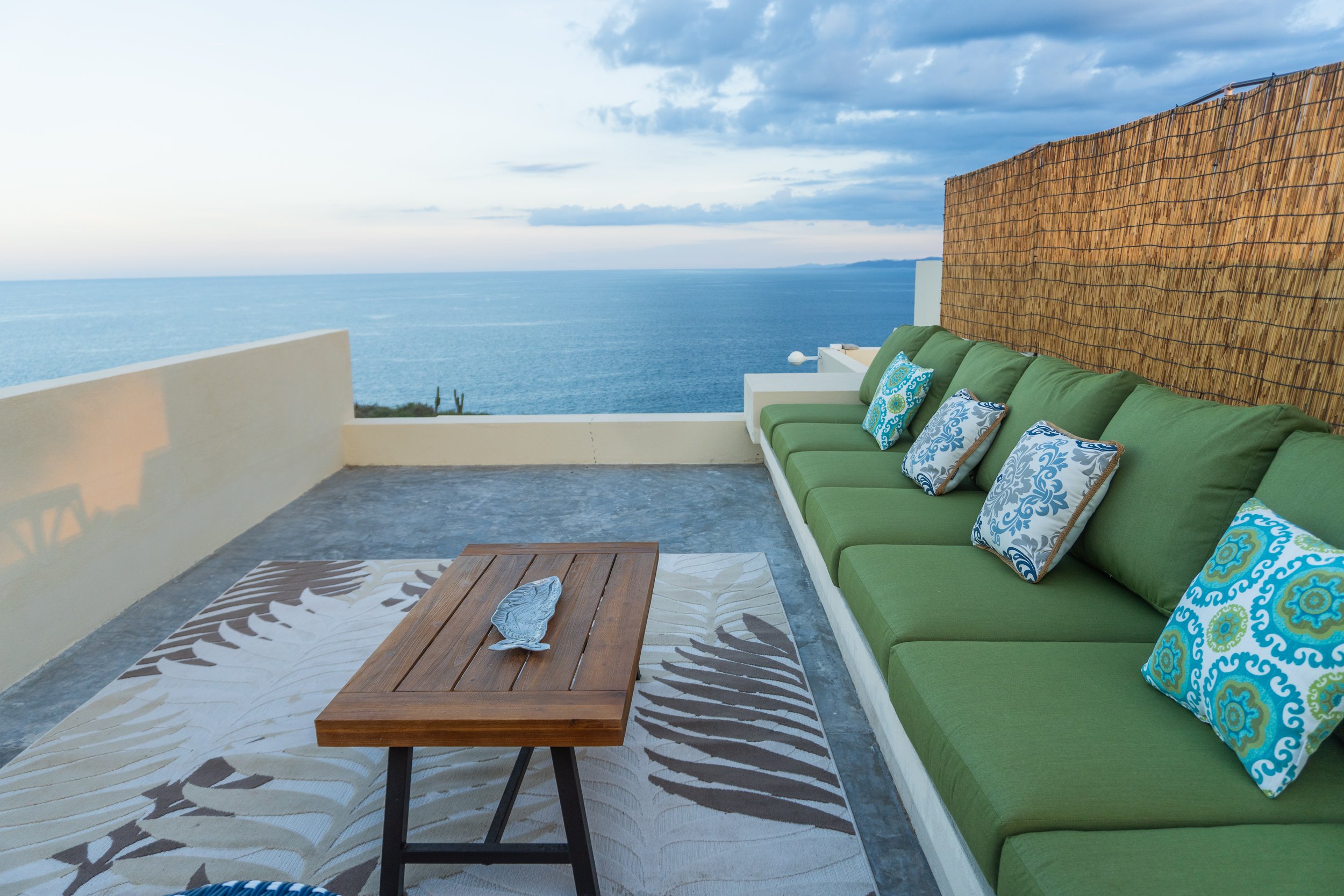 Outdoor terrace with green cushioned seating along the wall, decorative pillows, a wooden table, and a view of the ocean under a partly cloudy sky.
