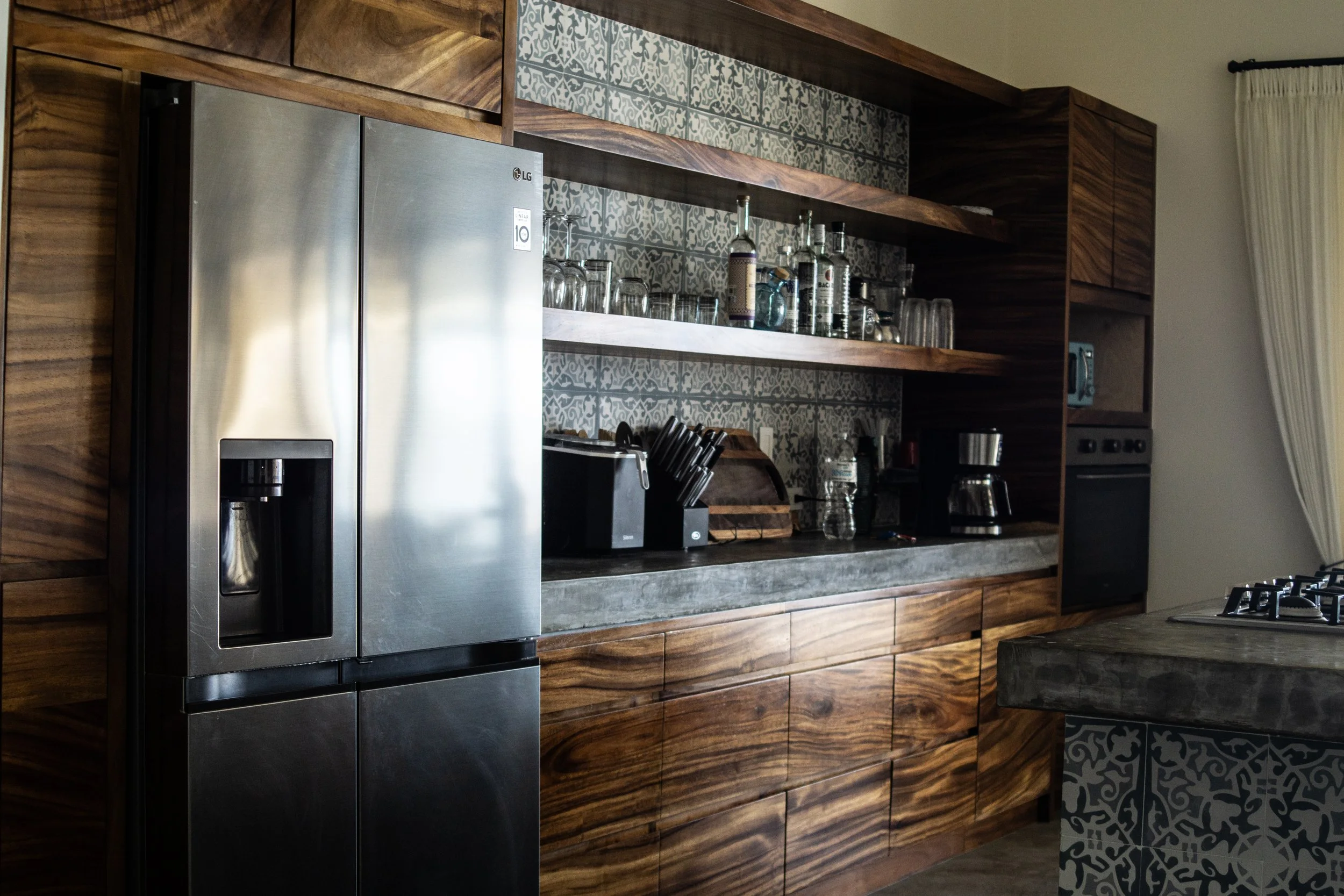 Modern kitchen with stainless steel refrigerator, wooden cabinets, open shelves with glasses and bottles, countertop with coffee maker and knives, patterned tiles on the wall and island.