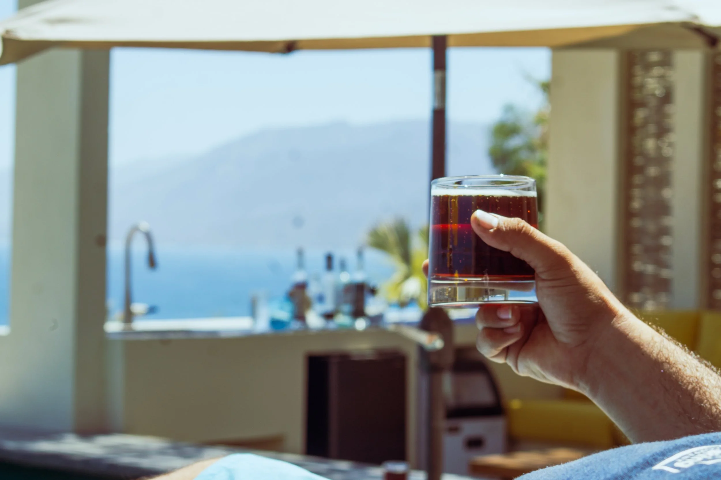 A person holding a glass of dark soda with ice, overlooking a scenic outdoor view of water and mountains.