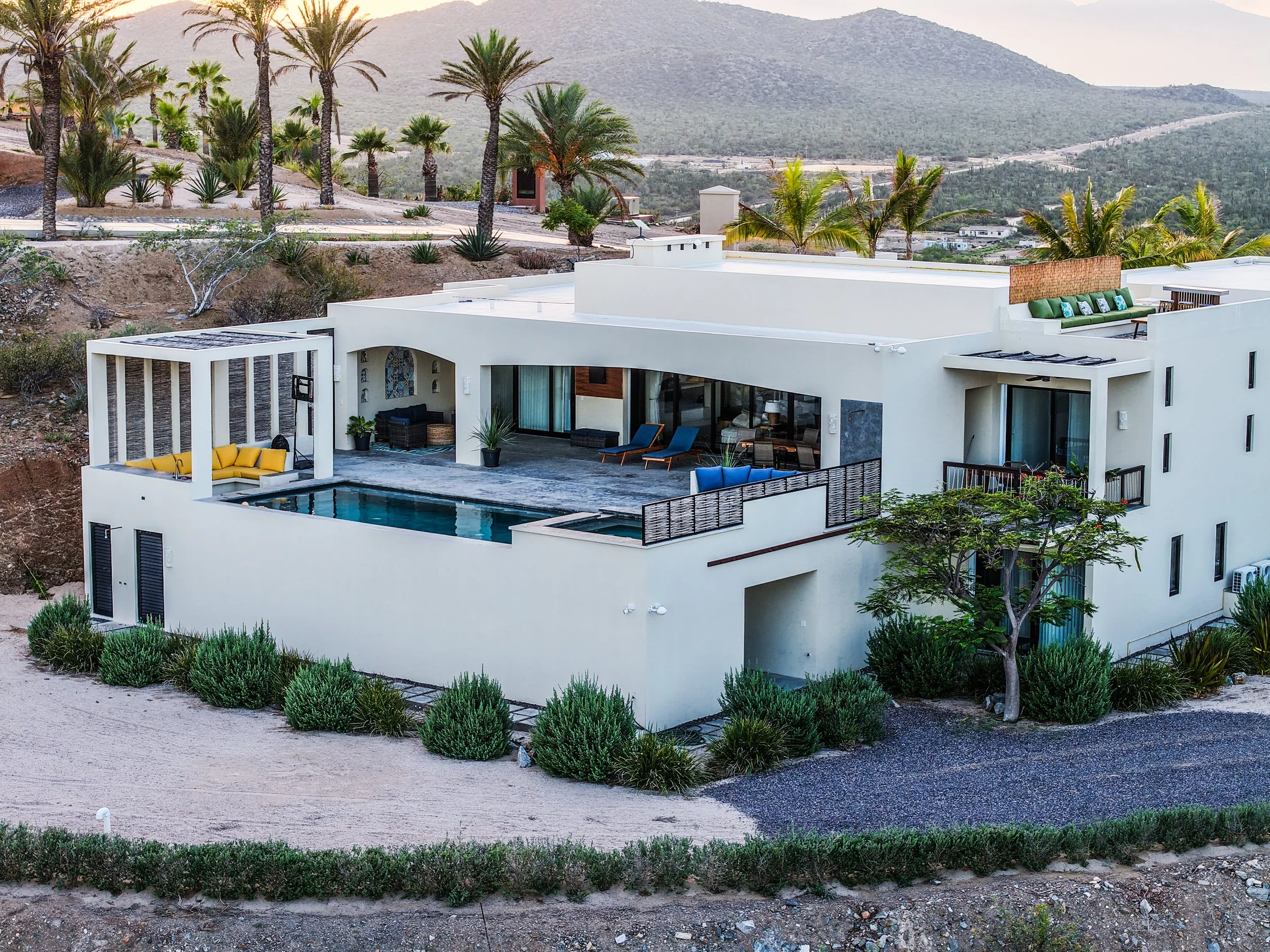 Modern white house with outdoor pool, surrounded by desert plants and palm trees, located in a hilly area with mountains in the background.