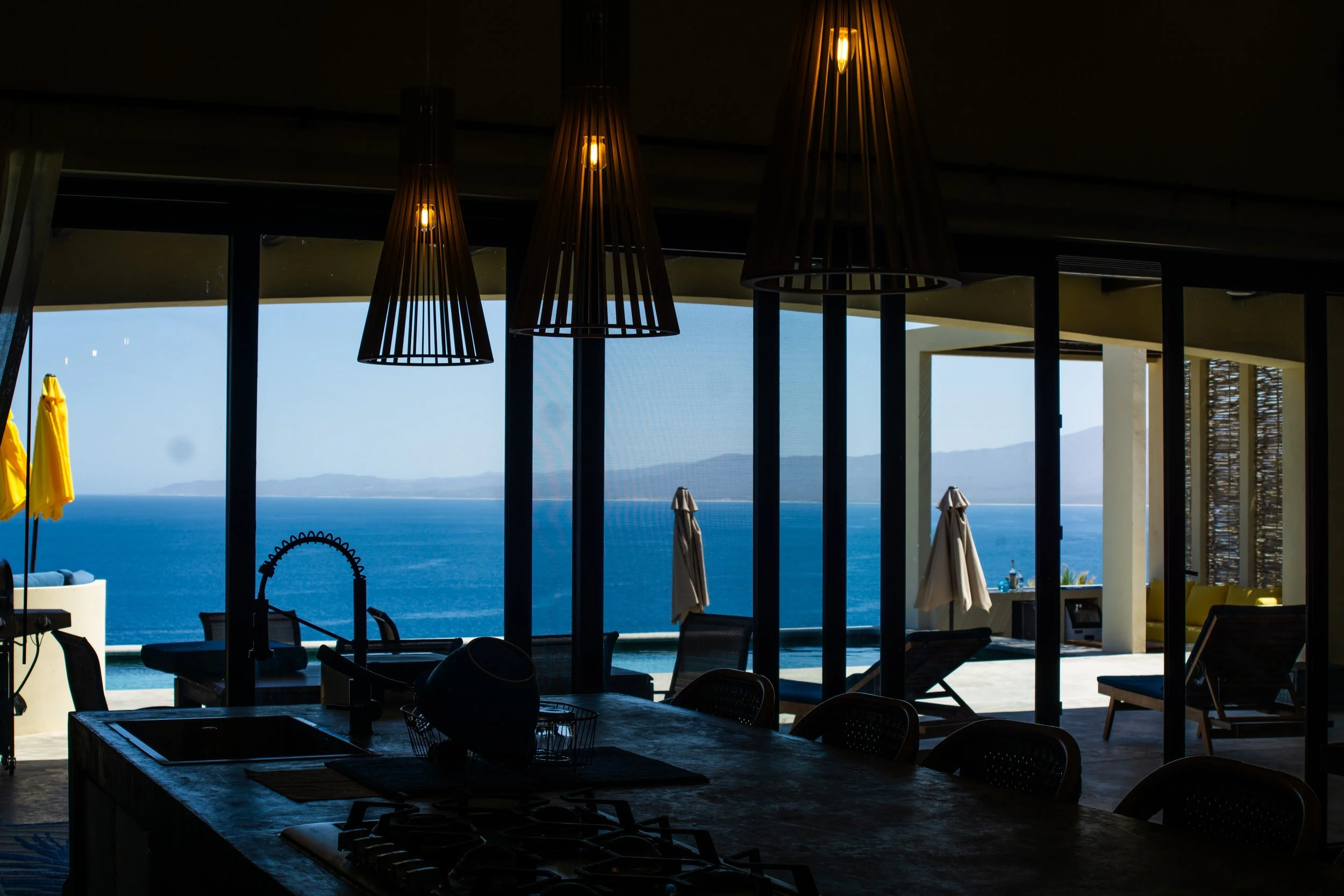 Interior view of a modern kitchen and living area with large glass windows overlooking the ocean, with hanging pendant lights, outdoor umbrellas, and chairs on a terrace.