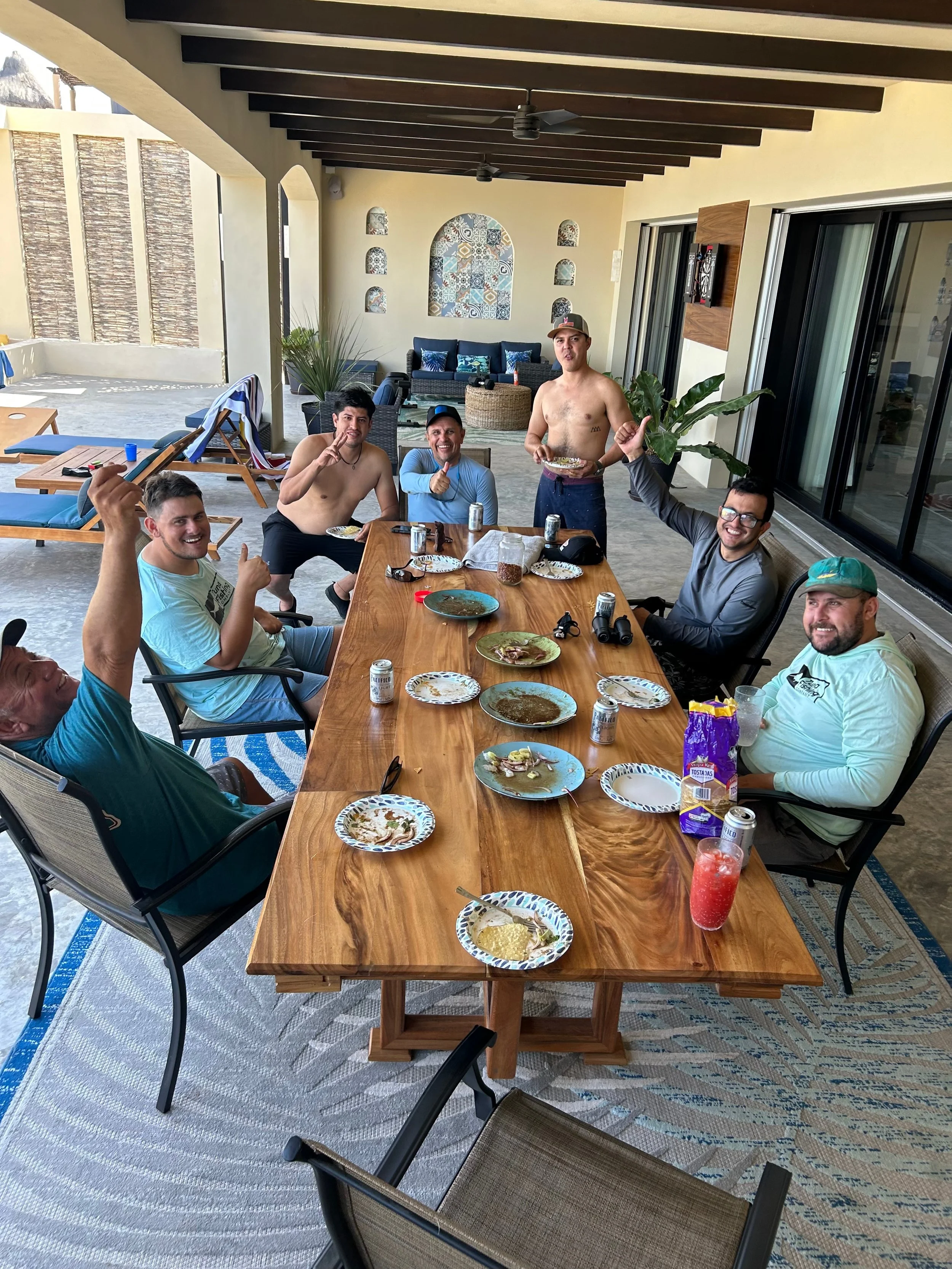 Group of seven men gathered around a large outdoor dining table on a patio, celebrating with some food and drinks, with some of them shirtless. The table is partially empty with a few plates, cans, and snacks remaining, and some men are smiling, giving thumbs-up, or holding food, indicating a casual, festive atmosphere.