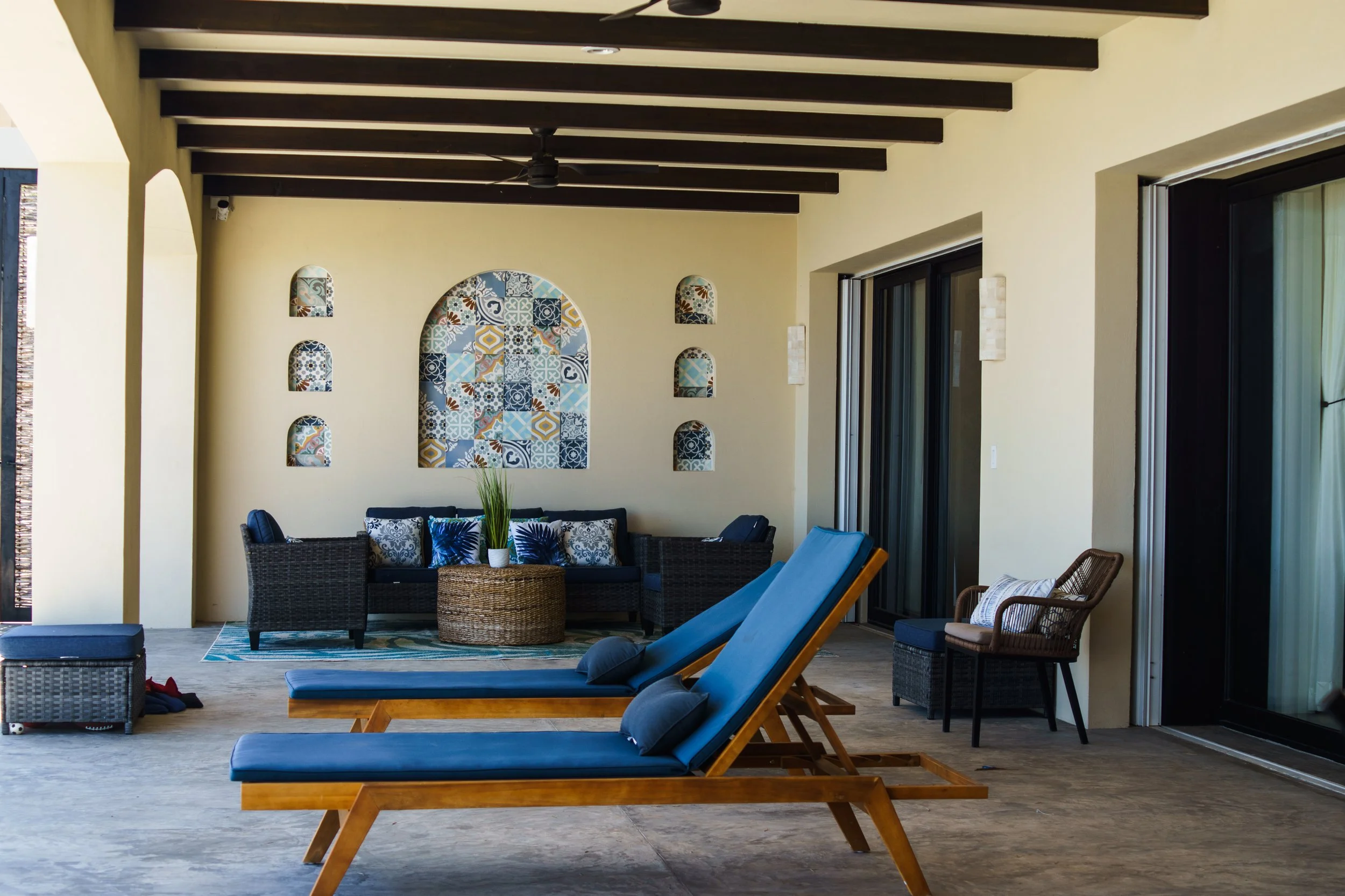 Covered outdoor patio with two blue lounge chairs, a black wicker sofa with blue and white pillows, a round wicker coffee table with a plant, a small wicker table with a chair, and decorative tiles on the wall. Large sliding glass doors on the right and ceiling fan overhead.
