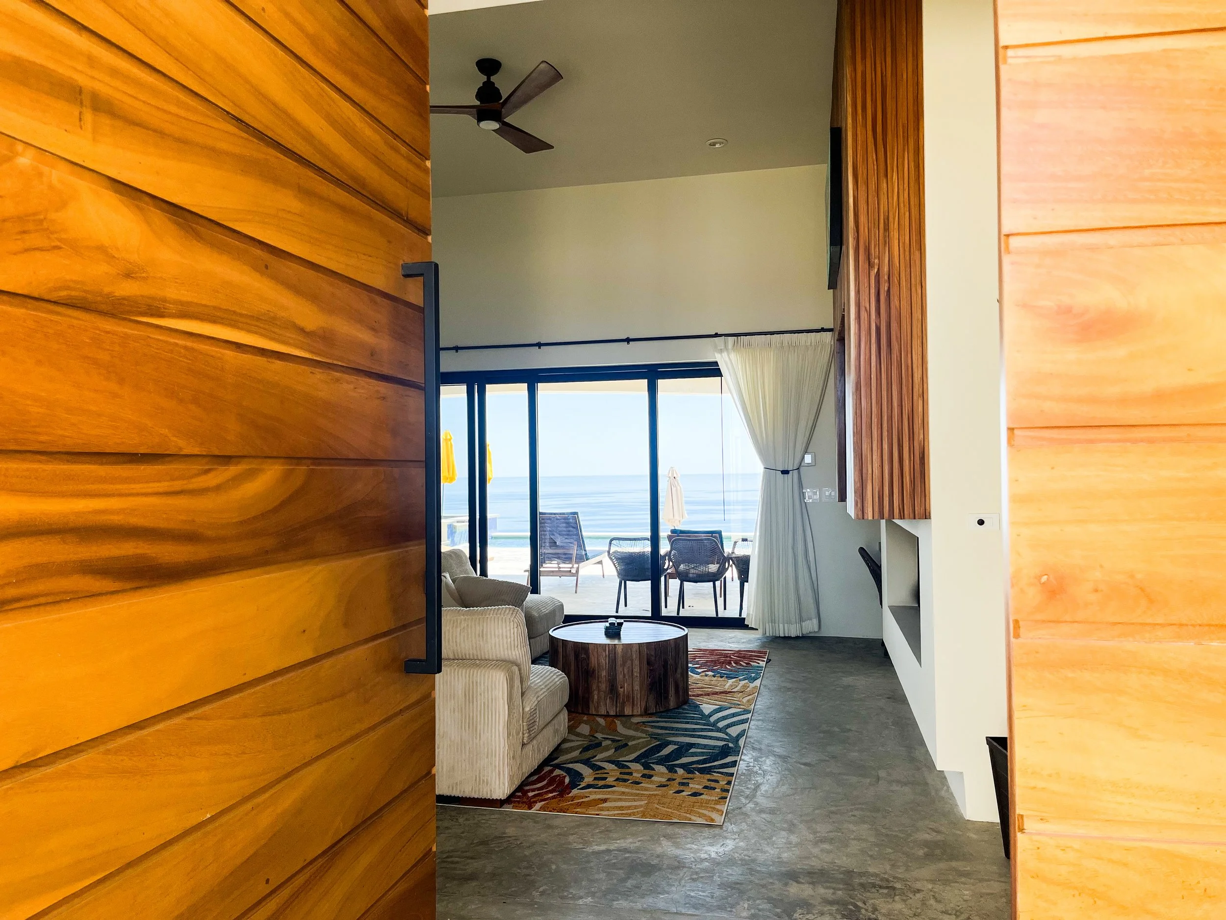 View of a living room with ocean view, featuring a sliding glass door, outdoor seating, an area rug, a beige armchair, a round wooden coffee table, and curtains.