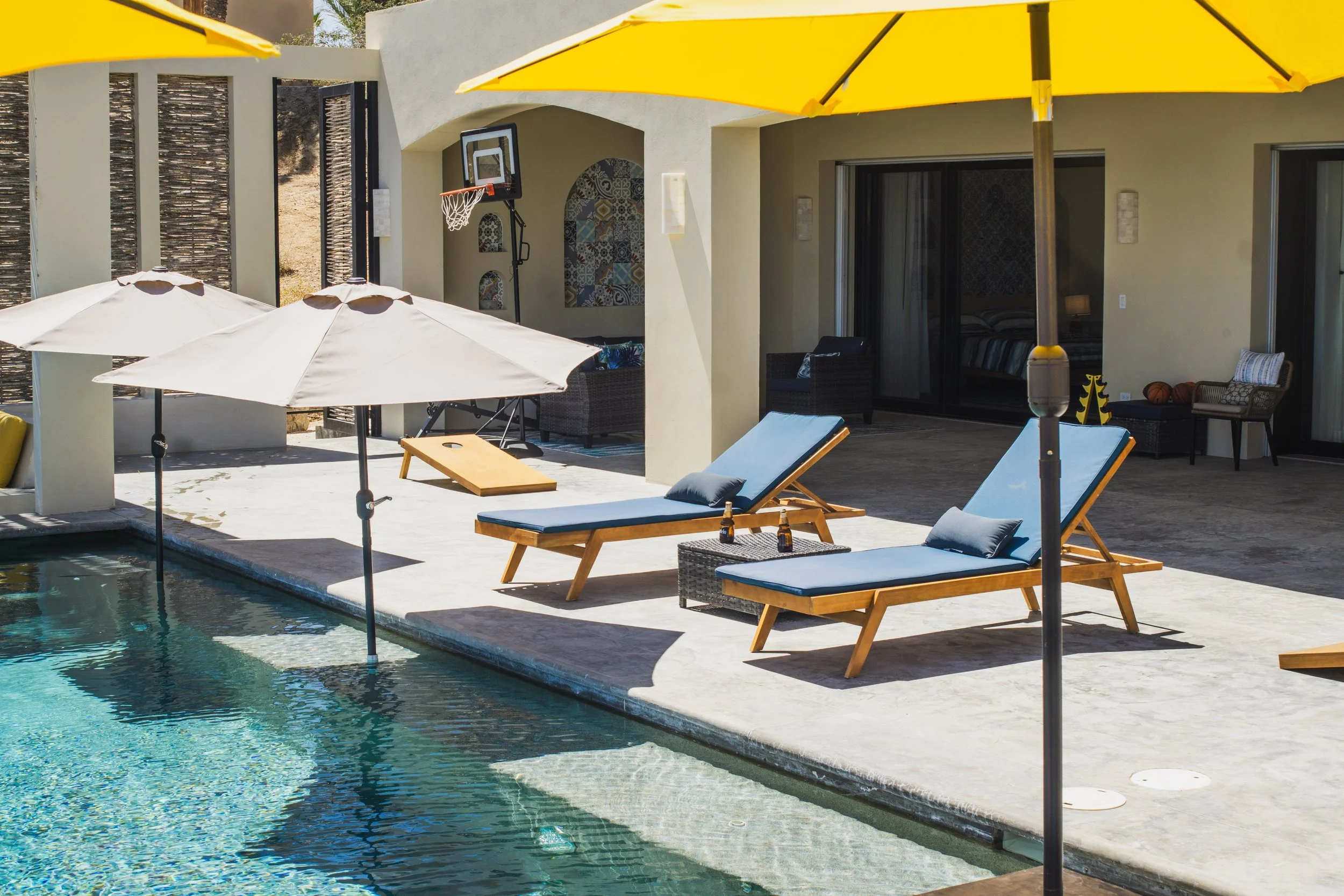 A poolside patio with two wooden lounge chairs with blue cushions, two umbrellas, and bottles on a small table. In the background, there are outdoor seating areas and a house with sliding glass doors.