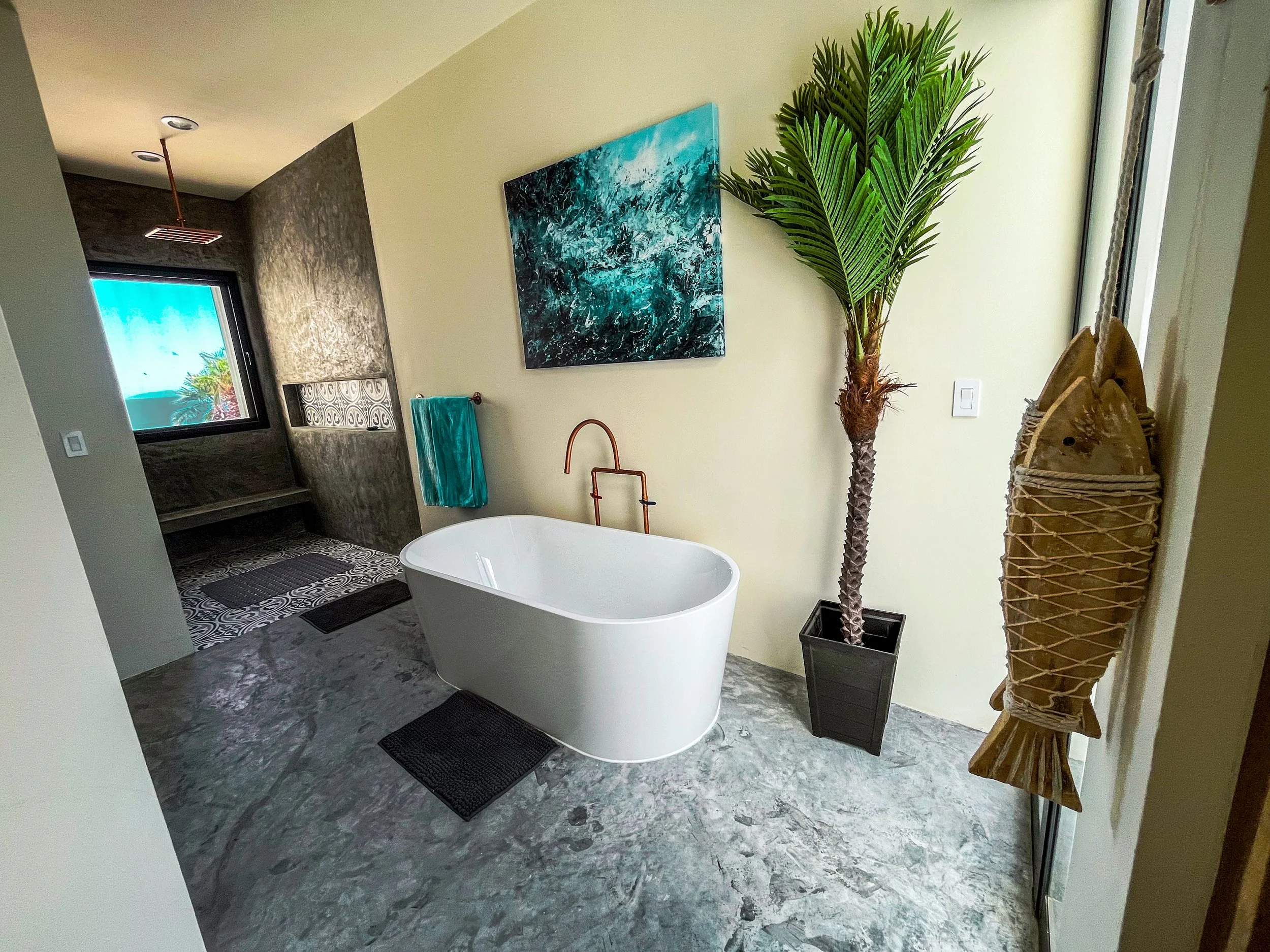 Modern bathroom with a freestanding bathtub, a large potted palm tree, an ocean-themed painting, boat decor, and a shower area with patterned wall and black and white tiles.