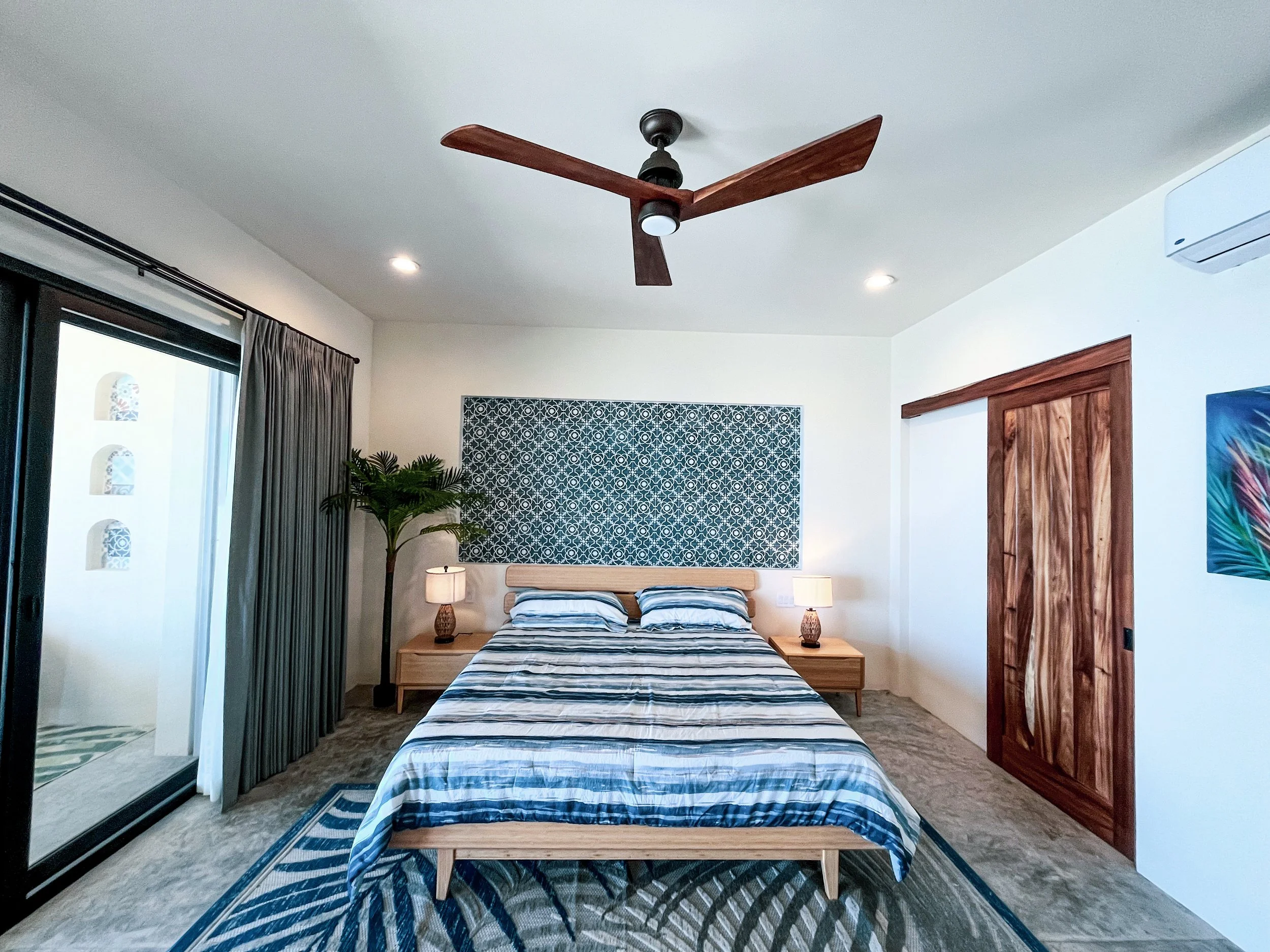 A modern bedroom with a wooden bed frame, striped bedding, two bedside lamps, a potted plant, and a decorative wall hanging above the bed. There is a sliding glass door with curtains on the left and a wooden sliding door on the right.