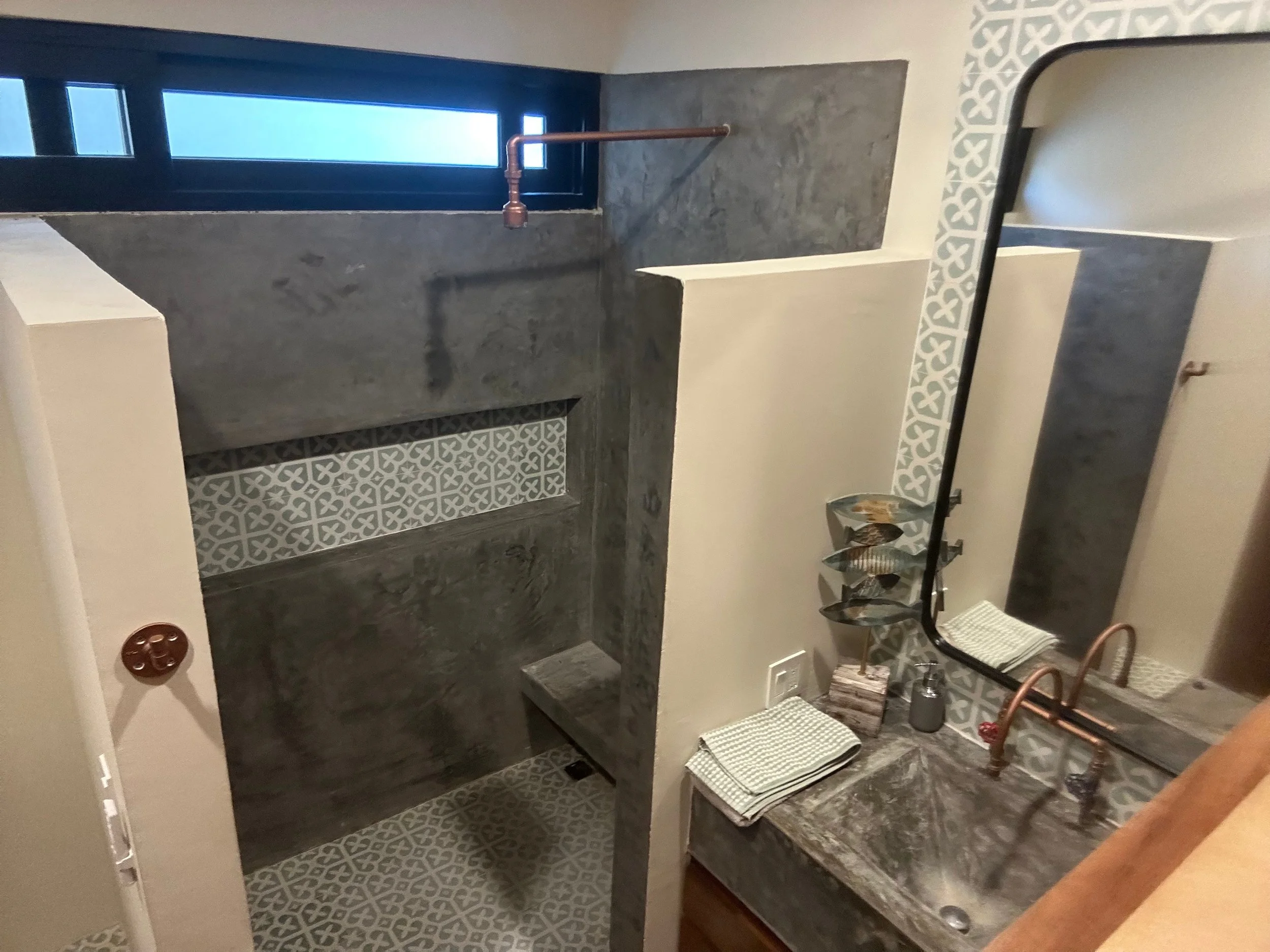 Bathroom with gray concrete shower area, patterned tiles, mirror, sink, copper faucet, and window.