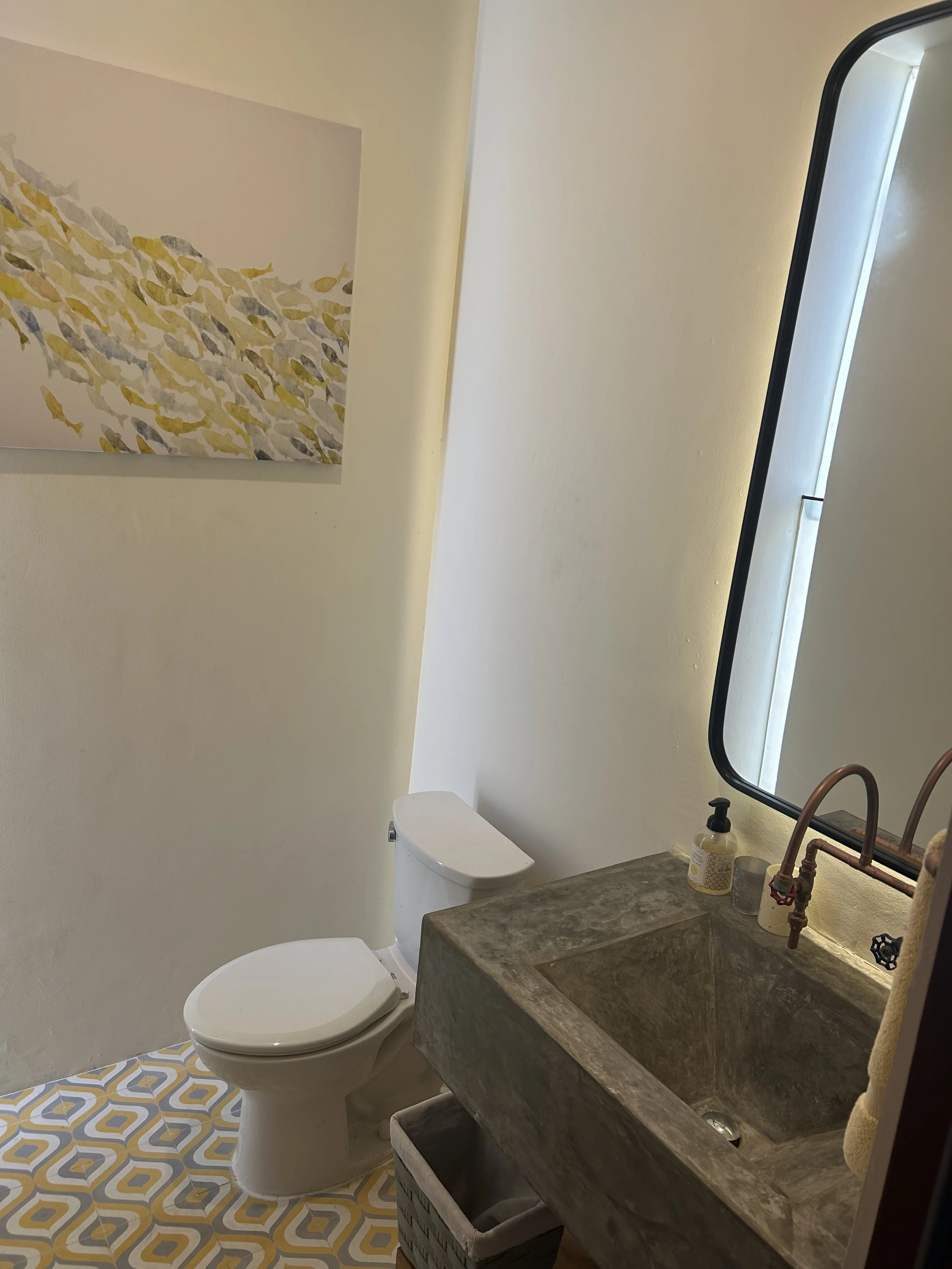A bathroom with a white toilet, a concrete sink with a wooden cabinet underneath, a large mirror, and a wall-mounted abstract painting. The floor has a yellow and gray geometric tile pattern.