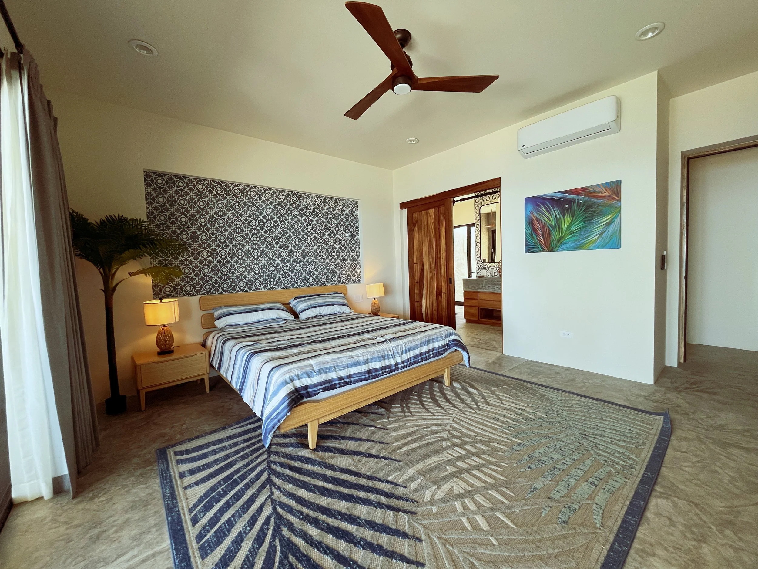 A bedroom with a wooden bed, striped bedding, two bedside lamps, a potted plant, a colorful abstract painting, air conditioning, and a wooden sliding door leading to a bathroom.
