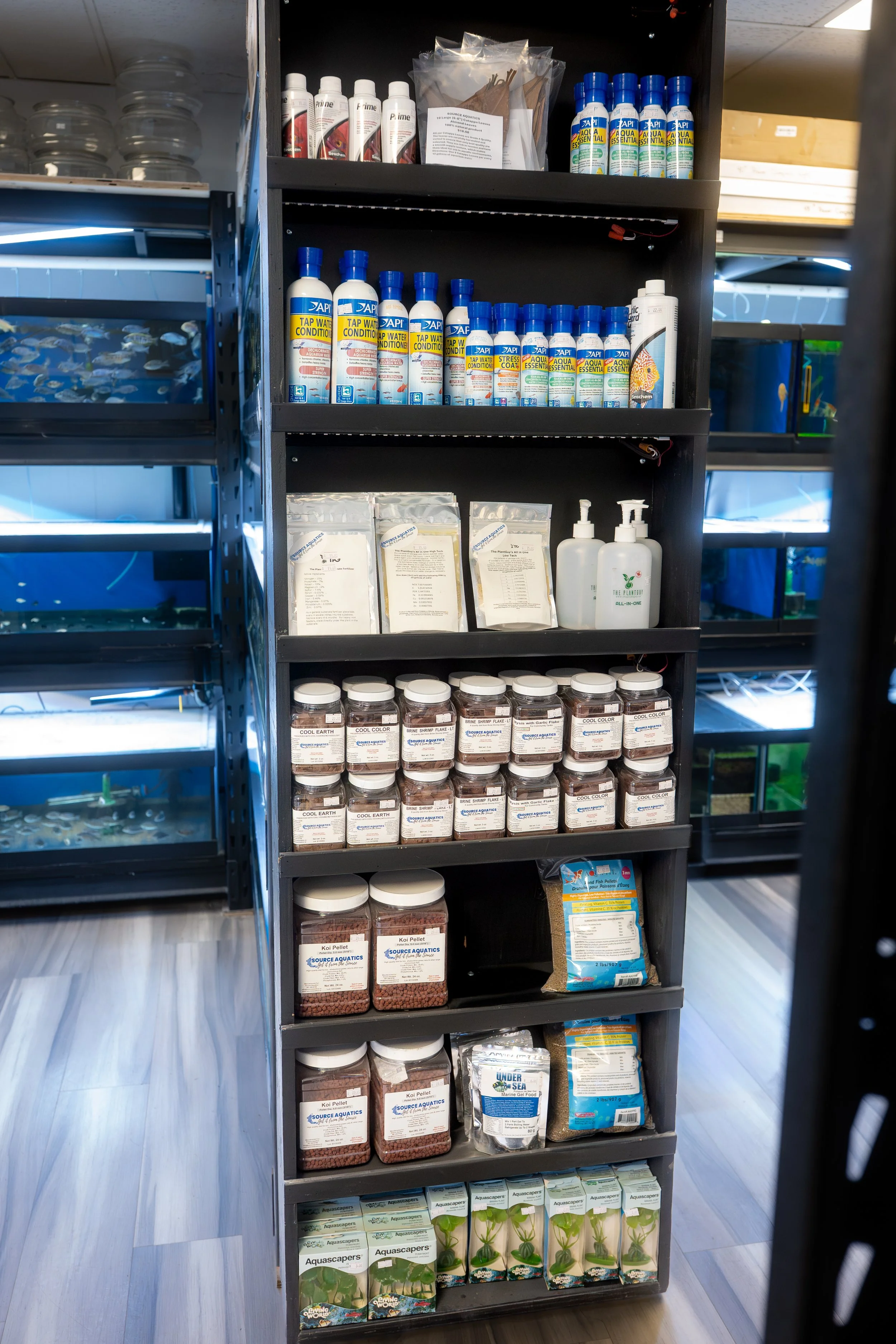 A black shelving unit in an aquarium store displaying various aquatic supplies, including water conditioners, some powders and liquids, fish food, and plant fertilizers.