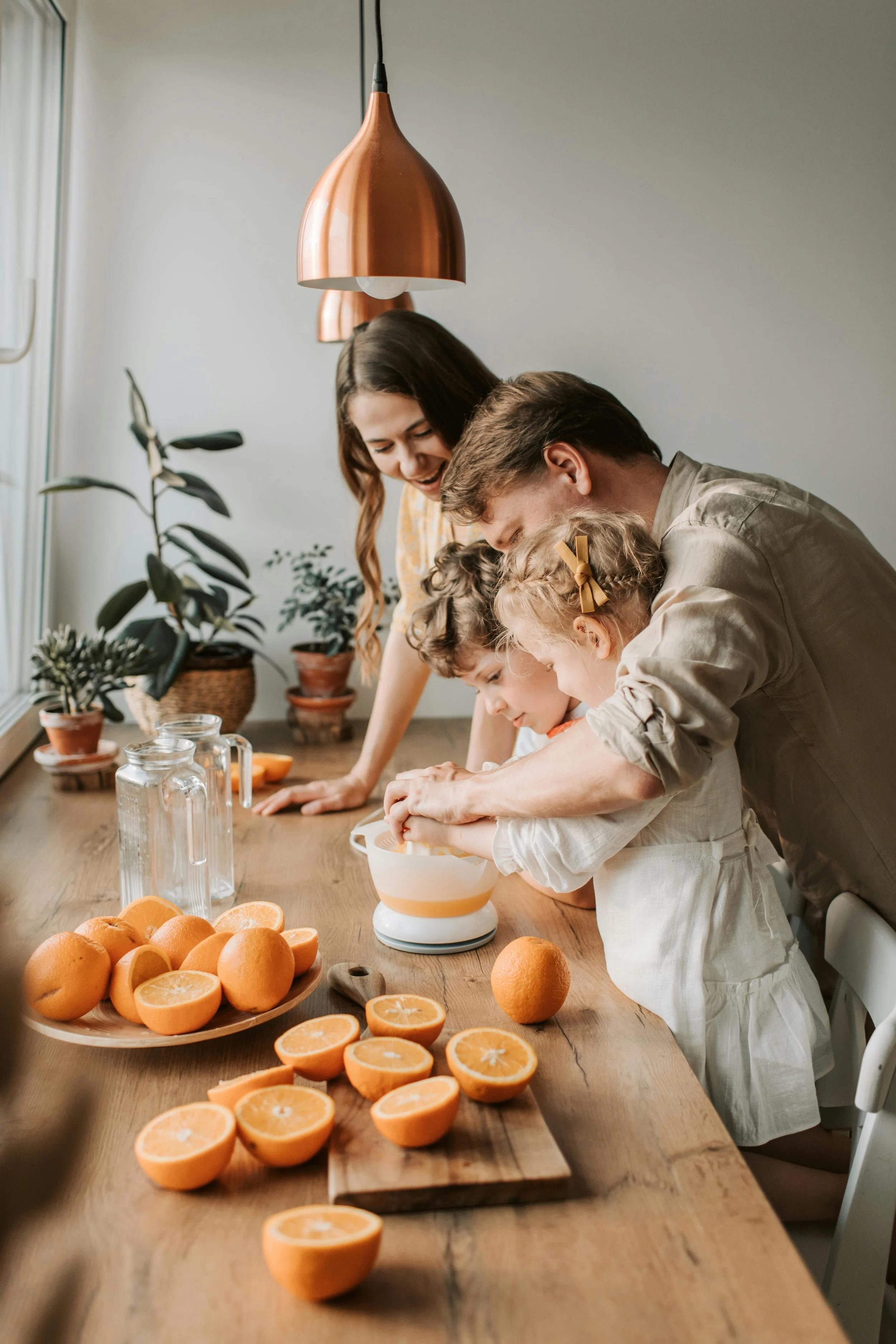 Family of four, including two young children, cooking together in the kitchen at a table with halved oranges, a bowl, and water bottles, smiling and enjoying the activity.