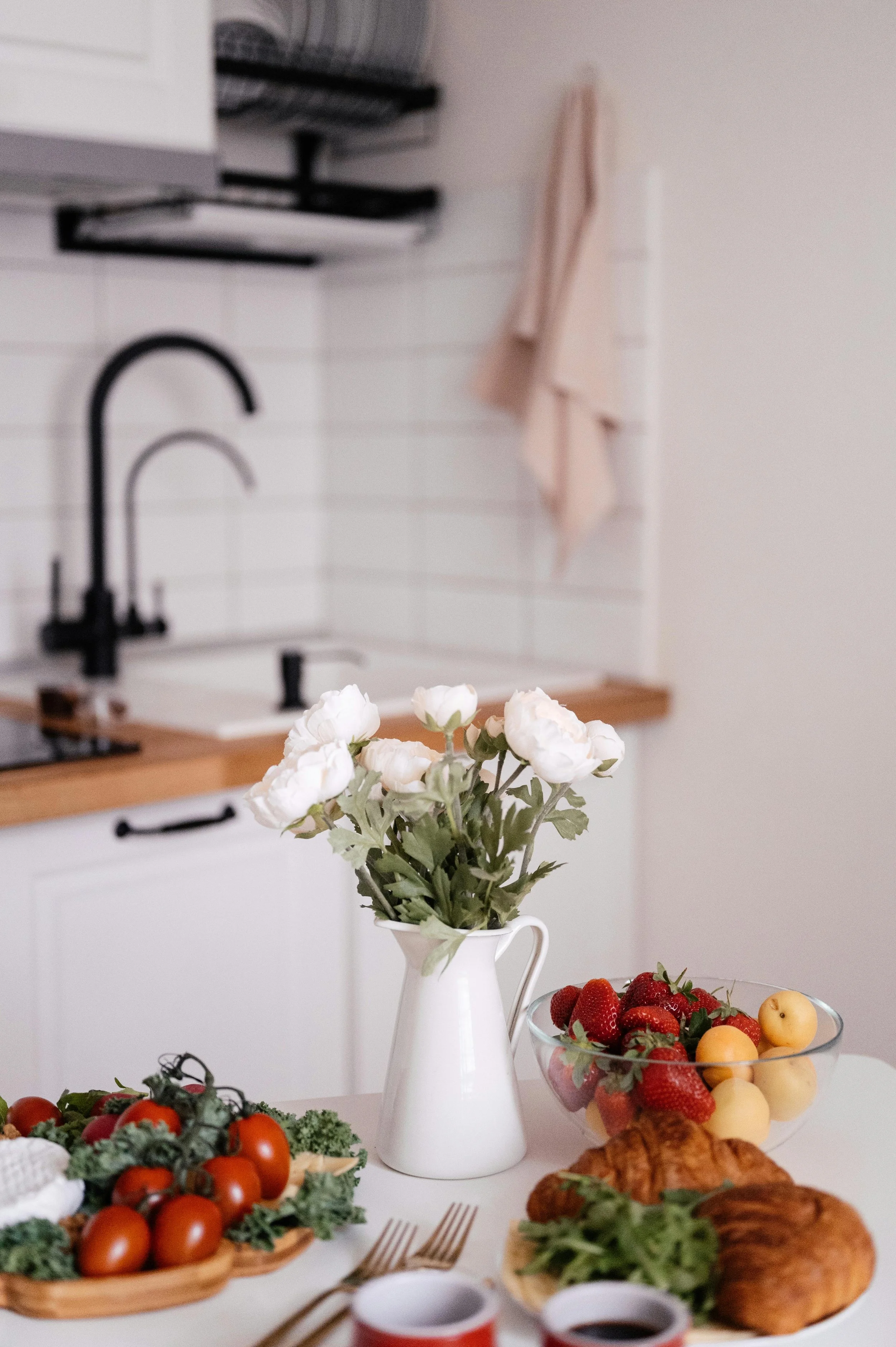 A cozy family kitchen scene with a vase of white flowers, fresh strawberries, yellow fruit, and croissants on the table, reflecting a pediatric dietitian’s realistic approach to balanced family meals.