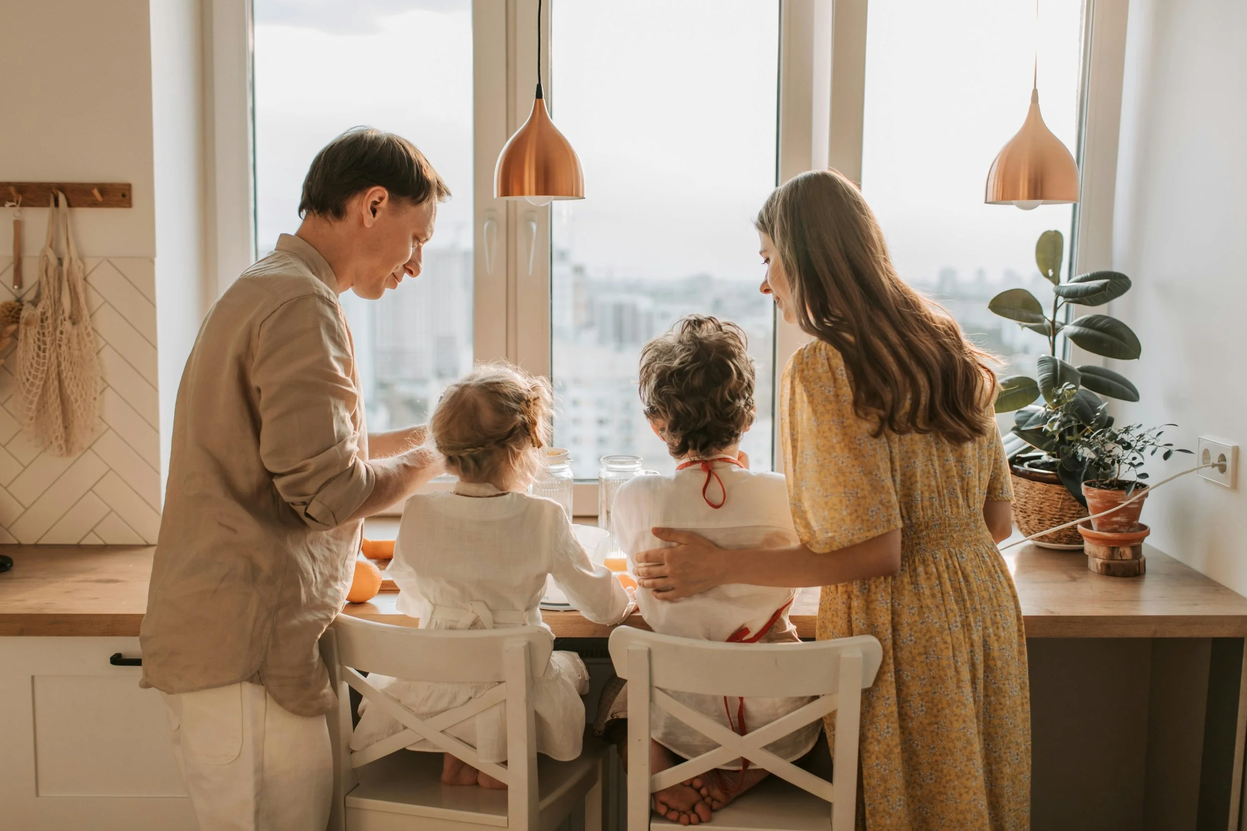 A family of four gathered in a bright, modern kitchen with children sitting on the countertops and plants by the window, reflecting a warm, real-life setting for family nutrition counseling and childrens nutrition support.