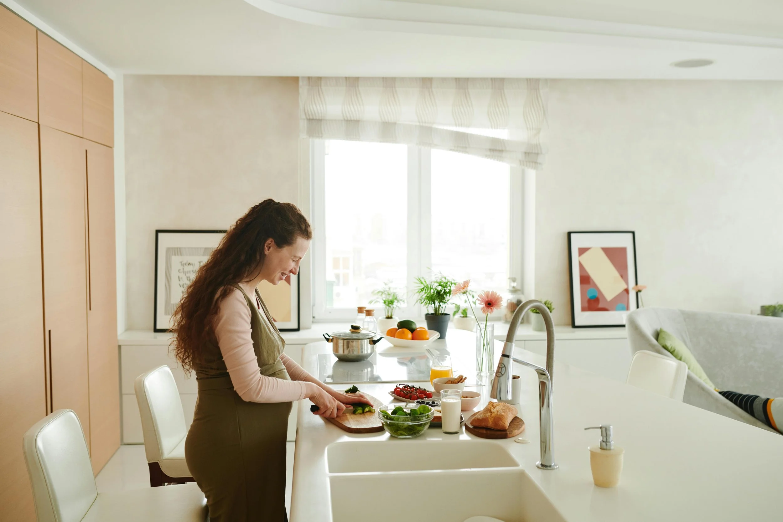 A woman with long curly hair preparing food at a white countertop in a bright kitchen with fresh ingredients, artwork, and plants, showcasing a pediatric nutritionist creating healthy, family-friendly meals.