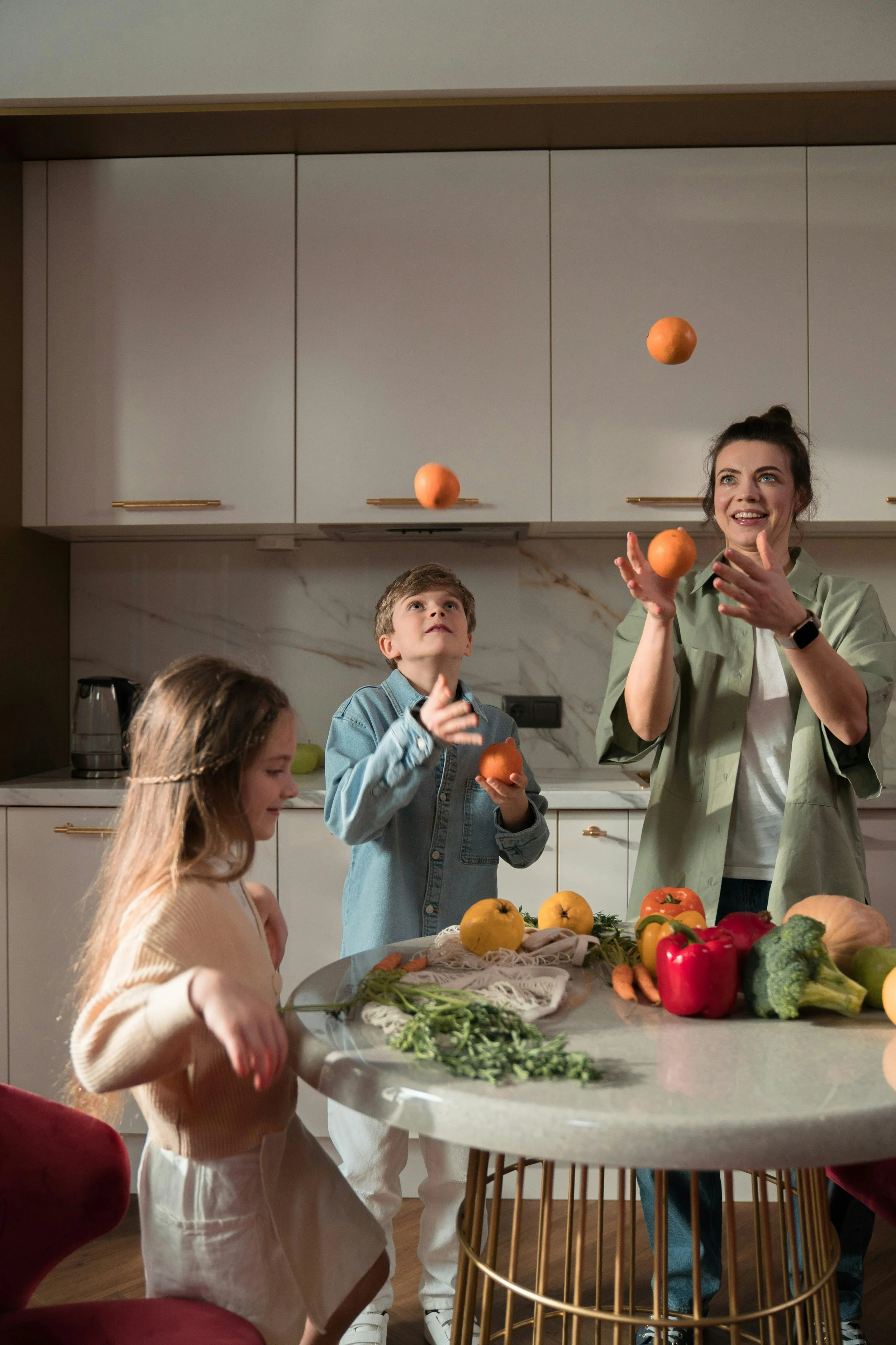A family nutritionist laughing with two children in a bright kitchen, tossing oranges in the air over a table filled with colorful fruits and vegetables, symbolizing fun, healthy eating for kids.