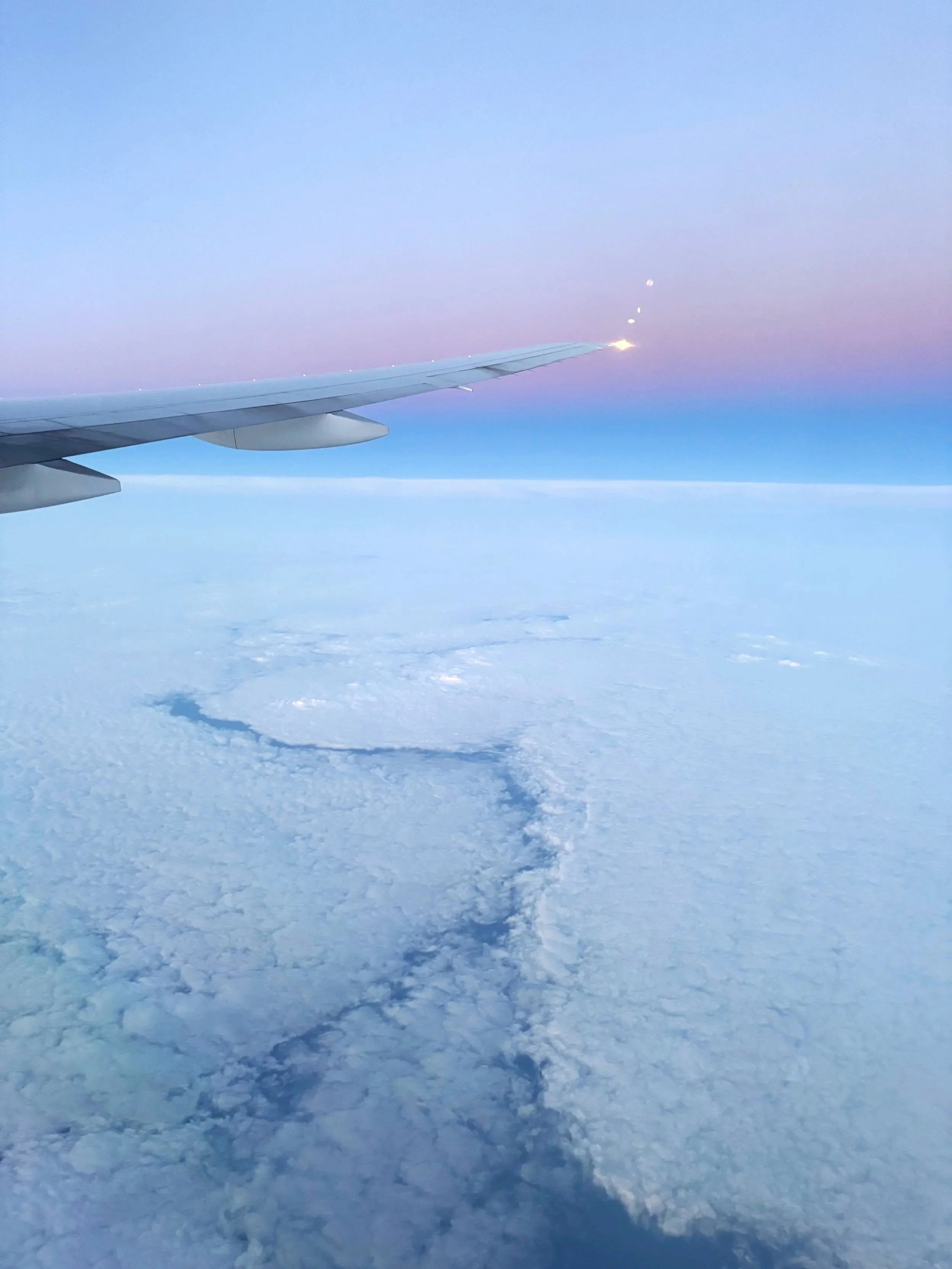 View from an airplane window showing wingtip, sky with clouds, and horizon during sunset or sunrise.