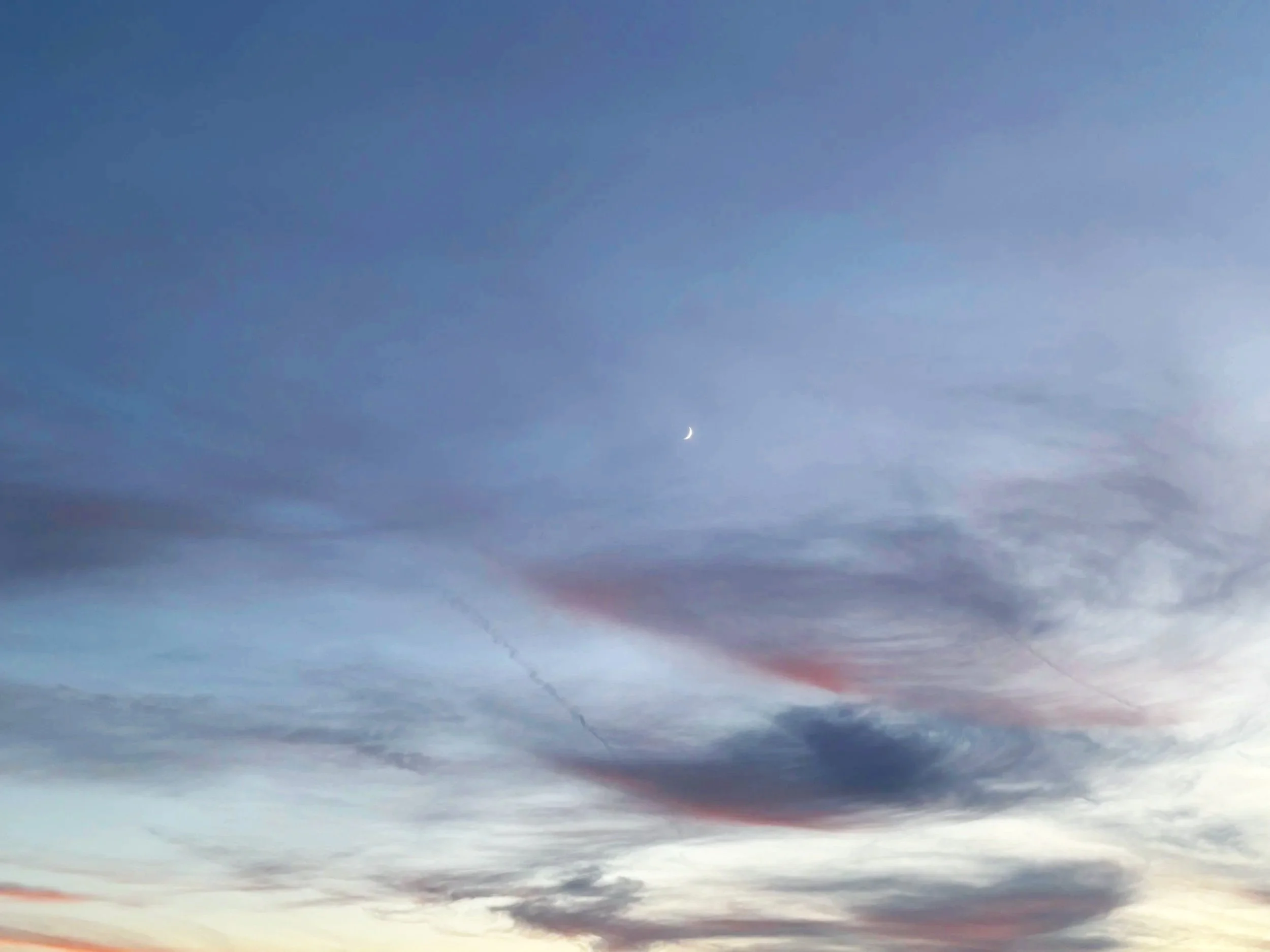 Sky with scattered clouds during sunset or sunrise, horizon showing a faint glow, and a crescent moon visible in the sky.