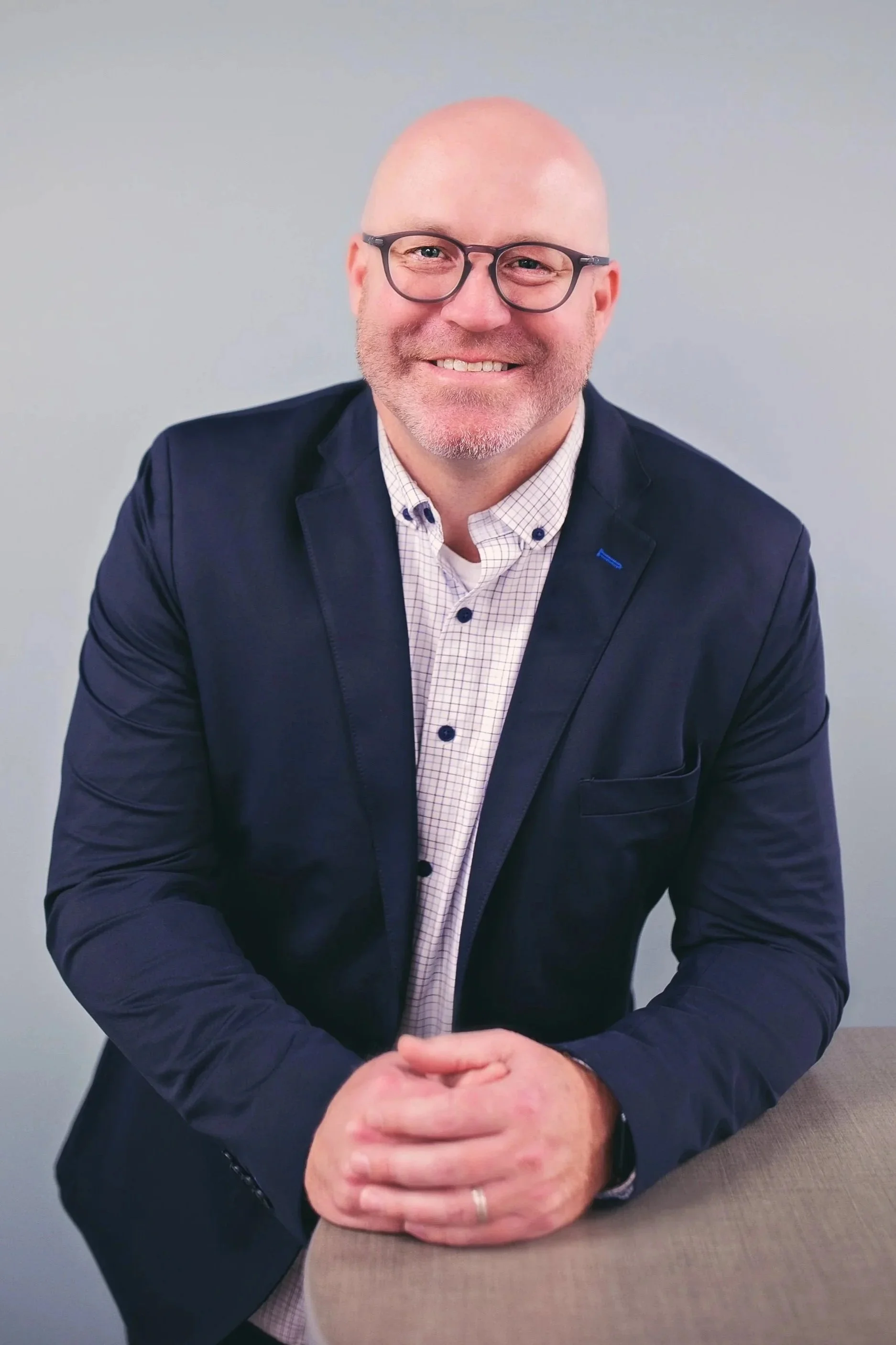 A smiling bald man with glasses, wearing a dark blazer over a checkered shirt, sitting with his hands clasped in front of him against a plain light gray background.