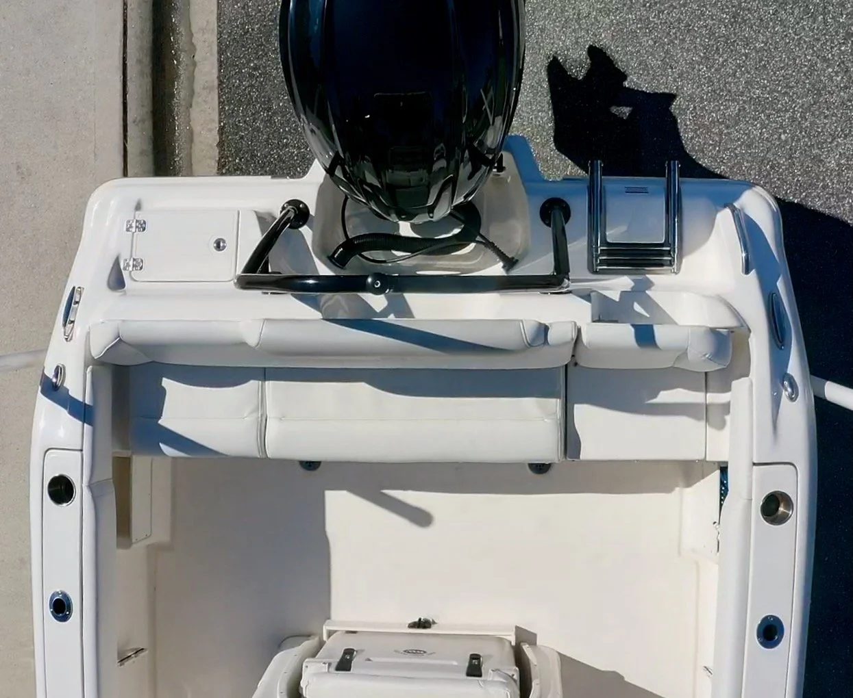 Top view of a small boat with a black outboard motor, white cushioned seat, ladder, and storage compartments on a paved surface.