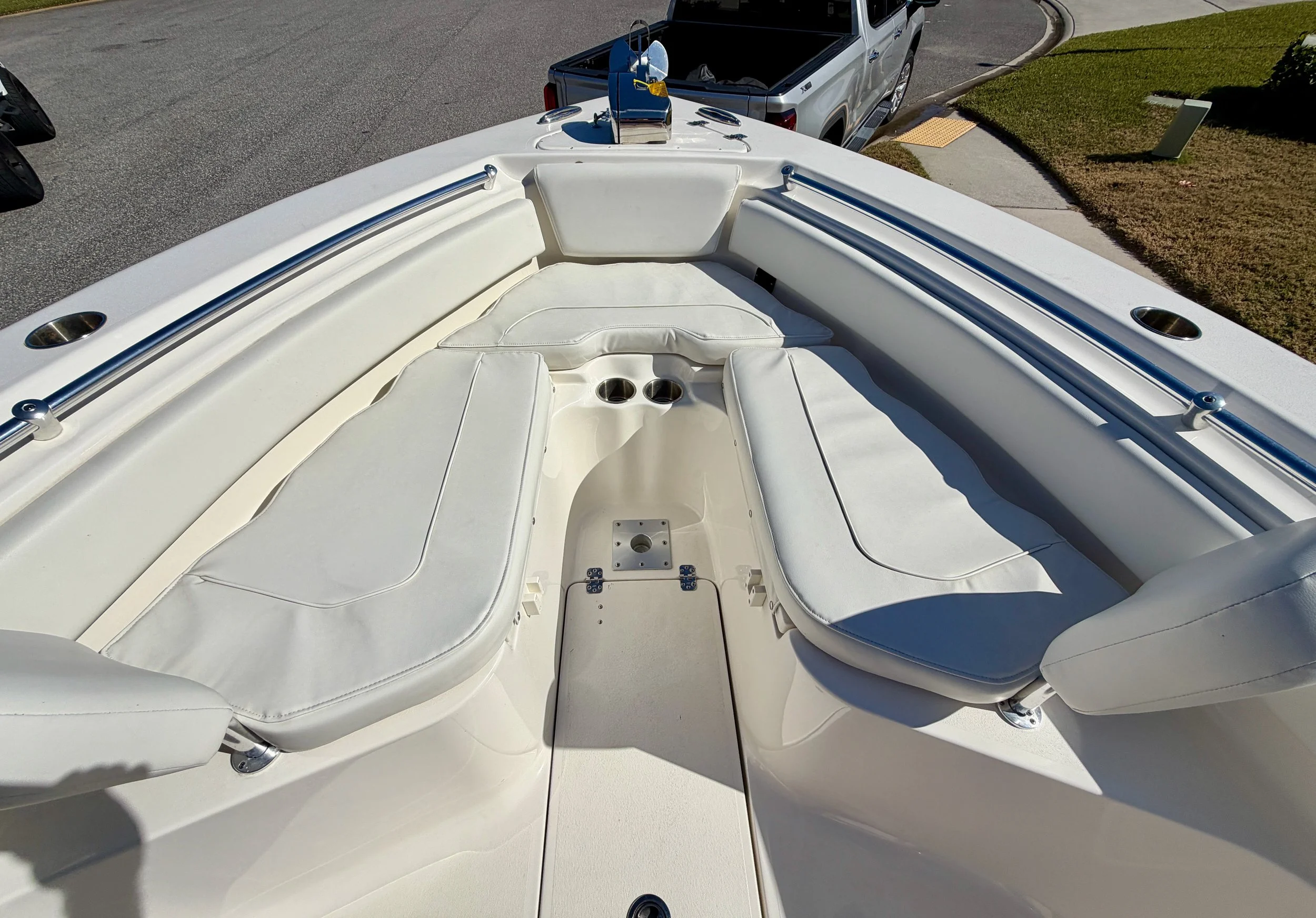 View of the bow seating area on a white boat, with cushioned seats and a small platform in the center, overlooking a parking lot and sidewalk.