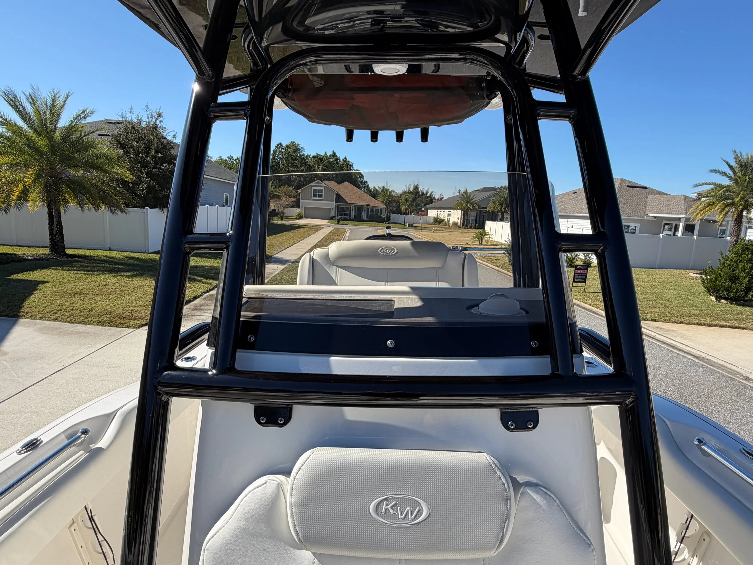 Inside view of a boat with a black T-top frame, white seating, and a windshield, on a suburban street with houses, palm trees, and a clear blue sky.
