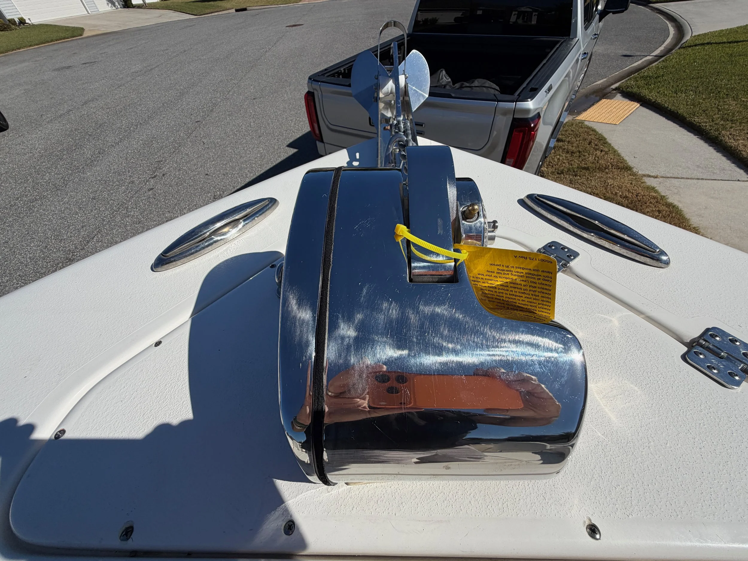 A boat's bow with a chrome anchor winch and winch cover, two oval chrome vents, and part of a boat trailer in background on a paved street.