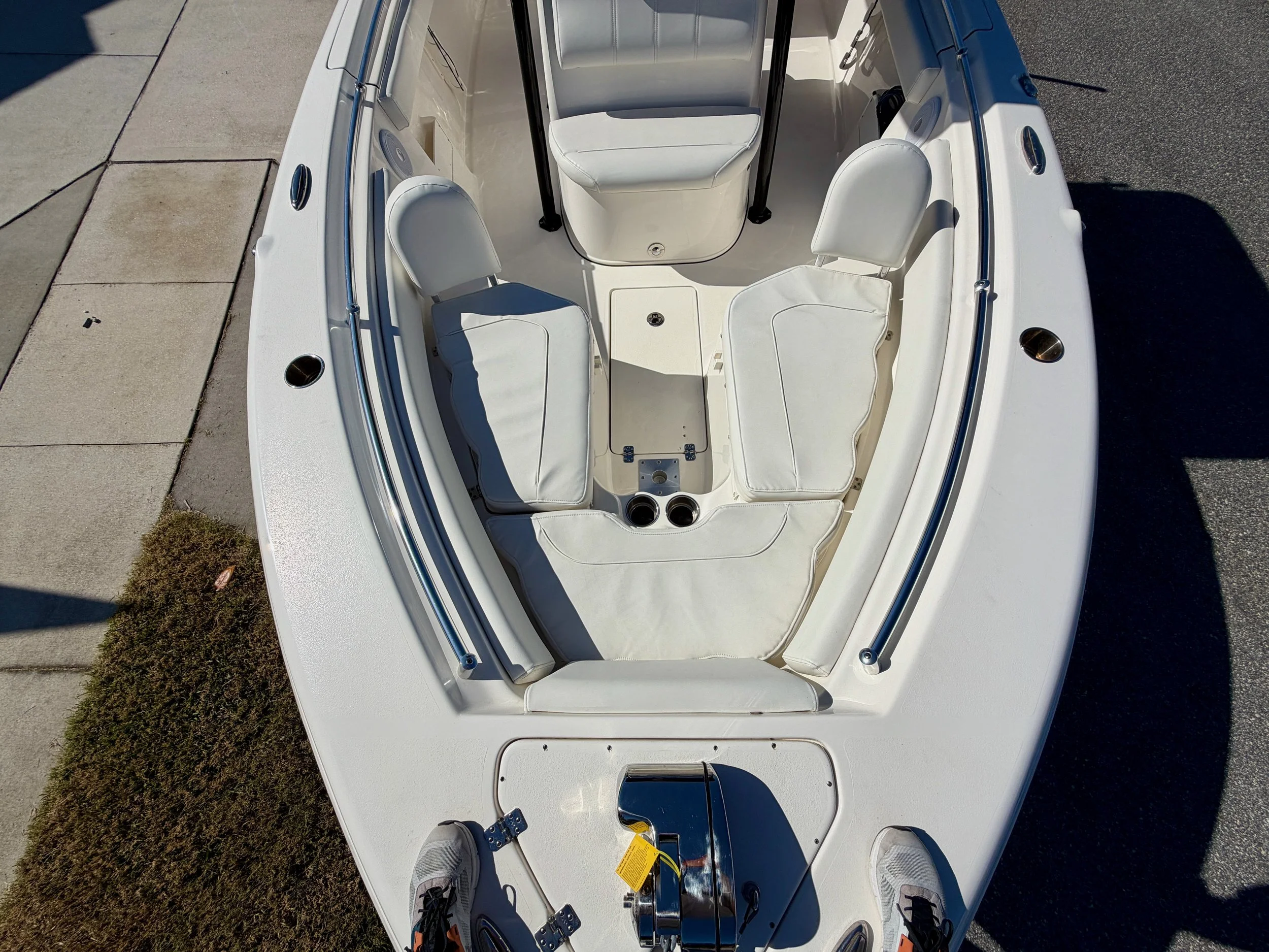 Top-down view of a white boat with padded seating inside, two cushioned seats at the front, a steering wheel, and a small hatch at the deck's center, with a person's feet visible at the bottom edge.