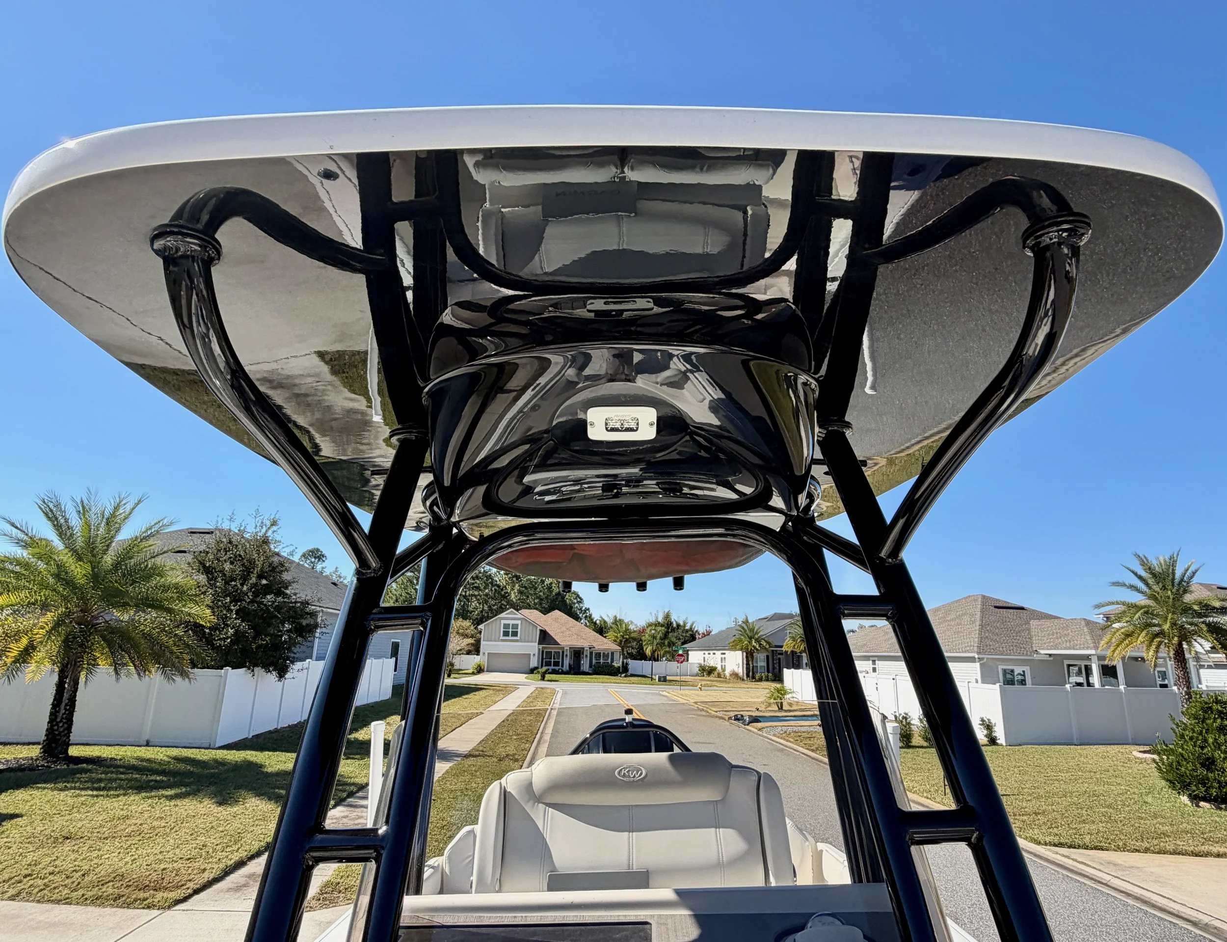 View of a boat's wakeboard tower from below, with a mirror and black hardware, overlooking a suburban street with houses and palm trees on a sunny day.