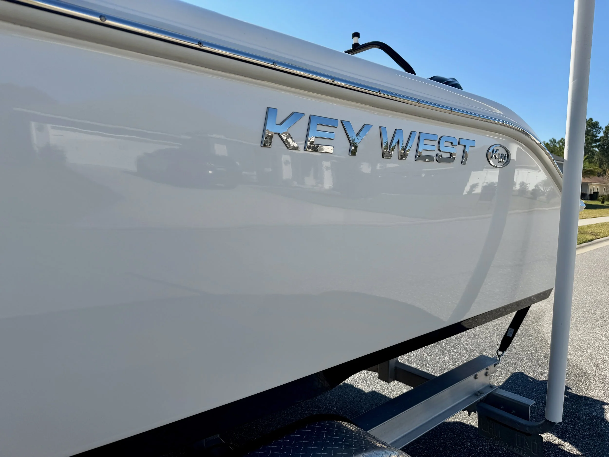 Close-up of a white boat with the brand name 'KEY WEST' in chrome letters on the side, partly visible against a background of a suburban neighborhood with green trees and houses under a blue sky.