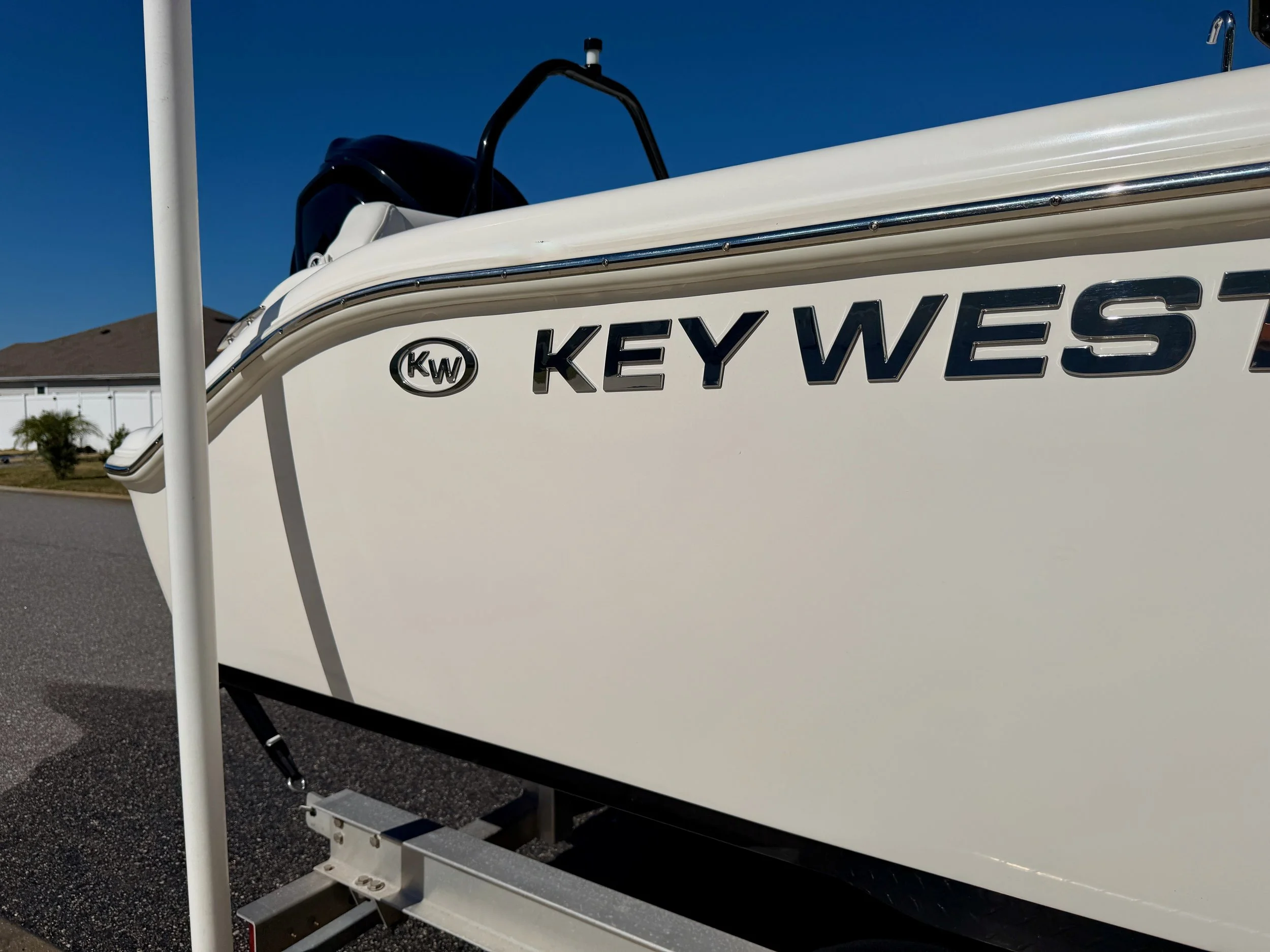 Close-up of a white boat with the label 'KEY WEST' and a logo with initials 'KW.' The boat is on a trailer, parked on a residential street with houses and trees in the background.