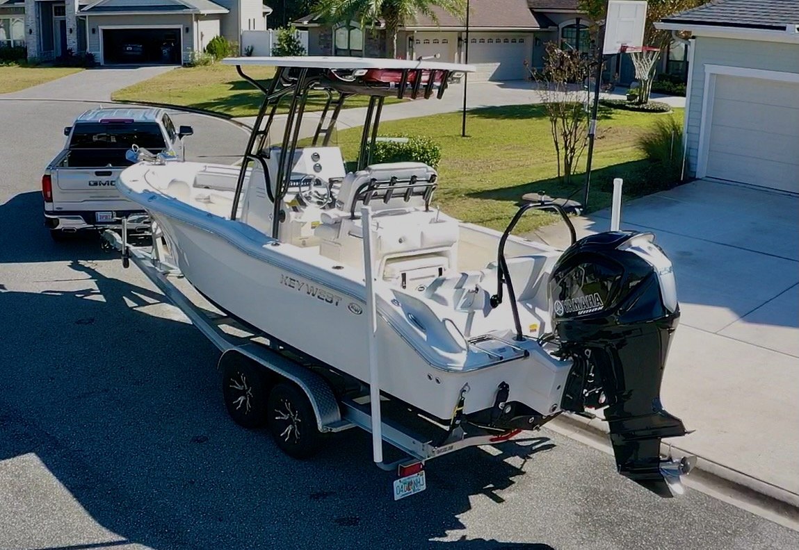 A white Key West boat on a trailer being towed by a GMC pickup truck in a suburban neighborhood driveway, with houses and a basketball hoop visible in the background.