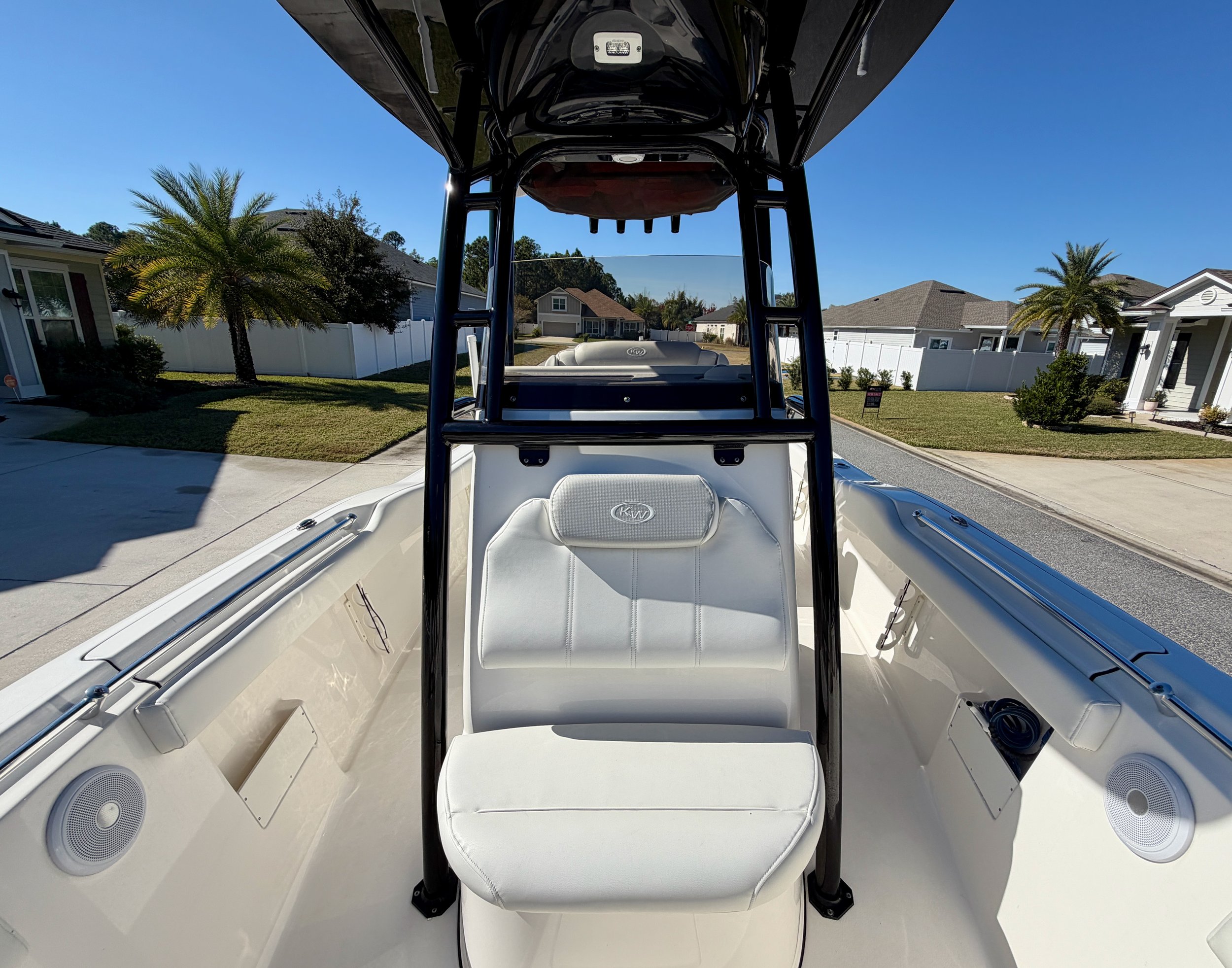 View of the bow of a white boat with a white padded seat and a black T-top, docked on a residential street with houses, palm trees, and a clear blue sky in the background.