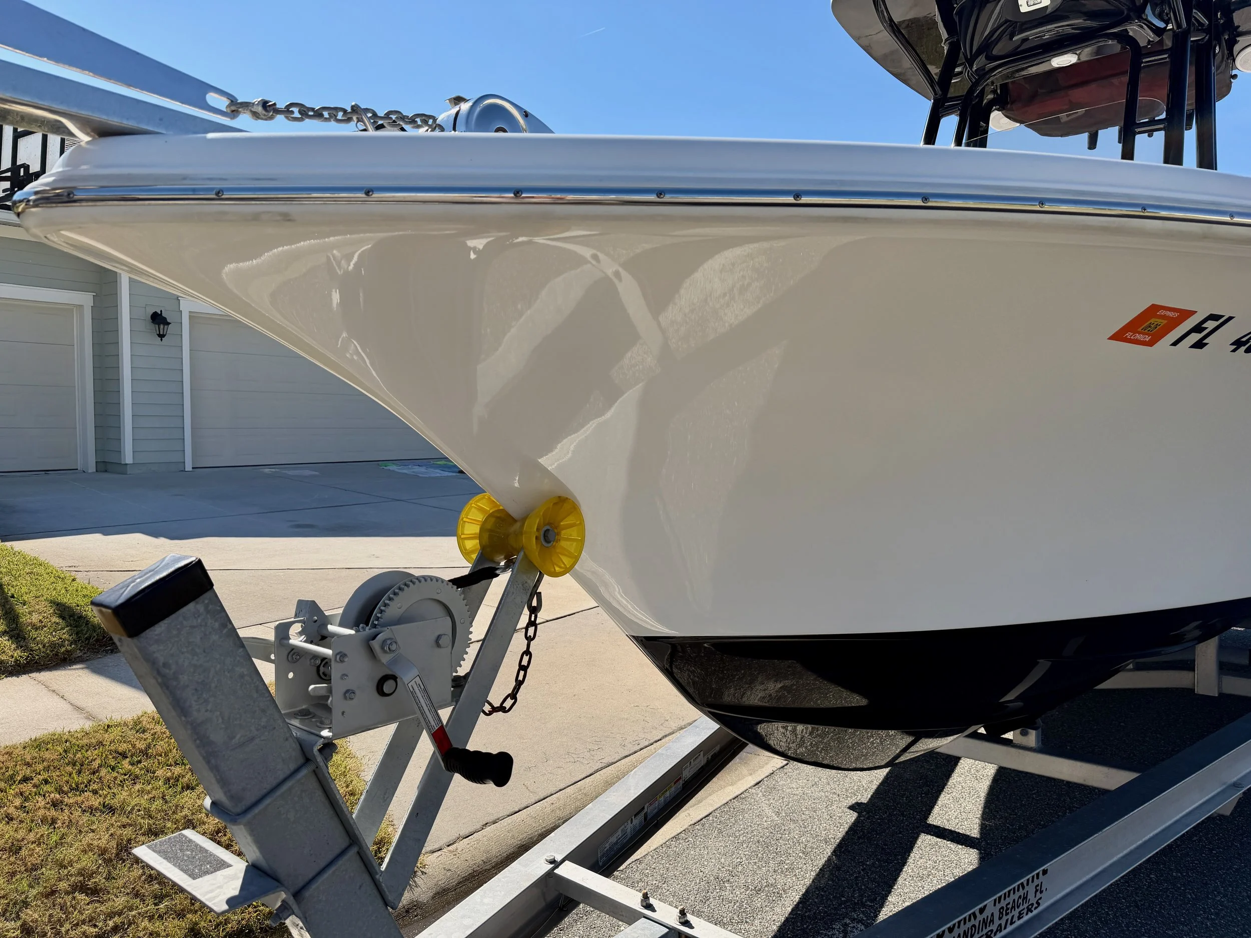 Close-up of a white boat on a trailer, with a yellow and gray winch and chain, parked on a driveway in front of a house.