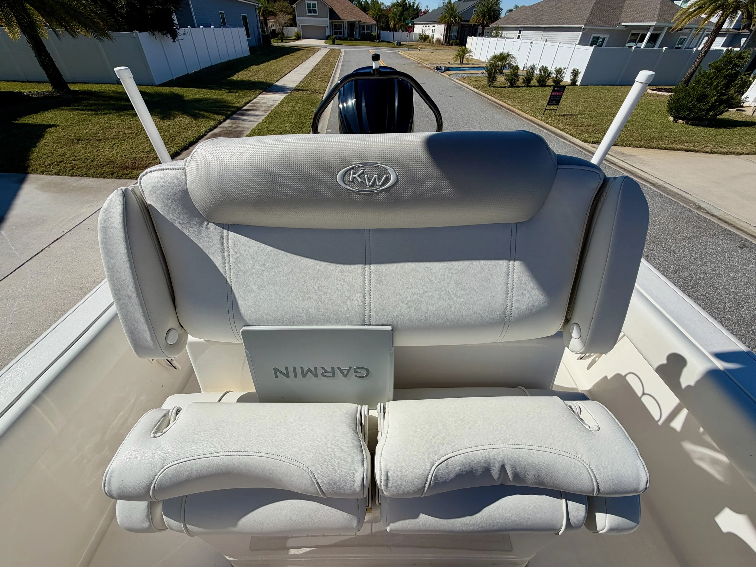 Photo of a white boat with white seats, a Garmin sign, and a black outboard motor, parked on a residential street with houses, lawns, trees, and a clear blue sky.