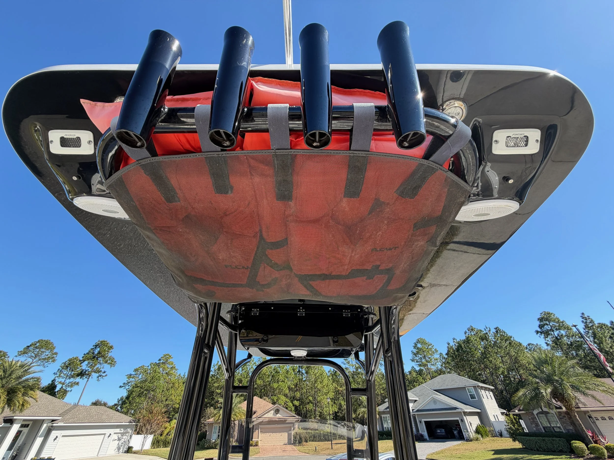 Looking up a boat with a black hull and orange life vest, parked upside down in a residential neighborhood with houses and trees under a clear blue sky.