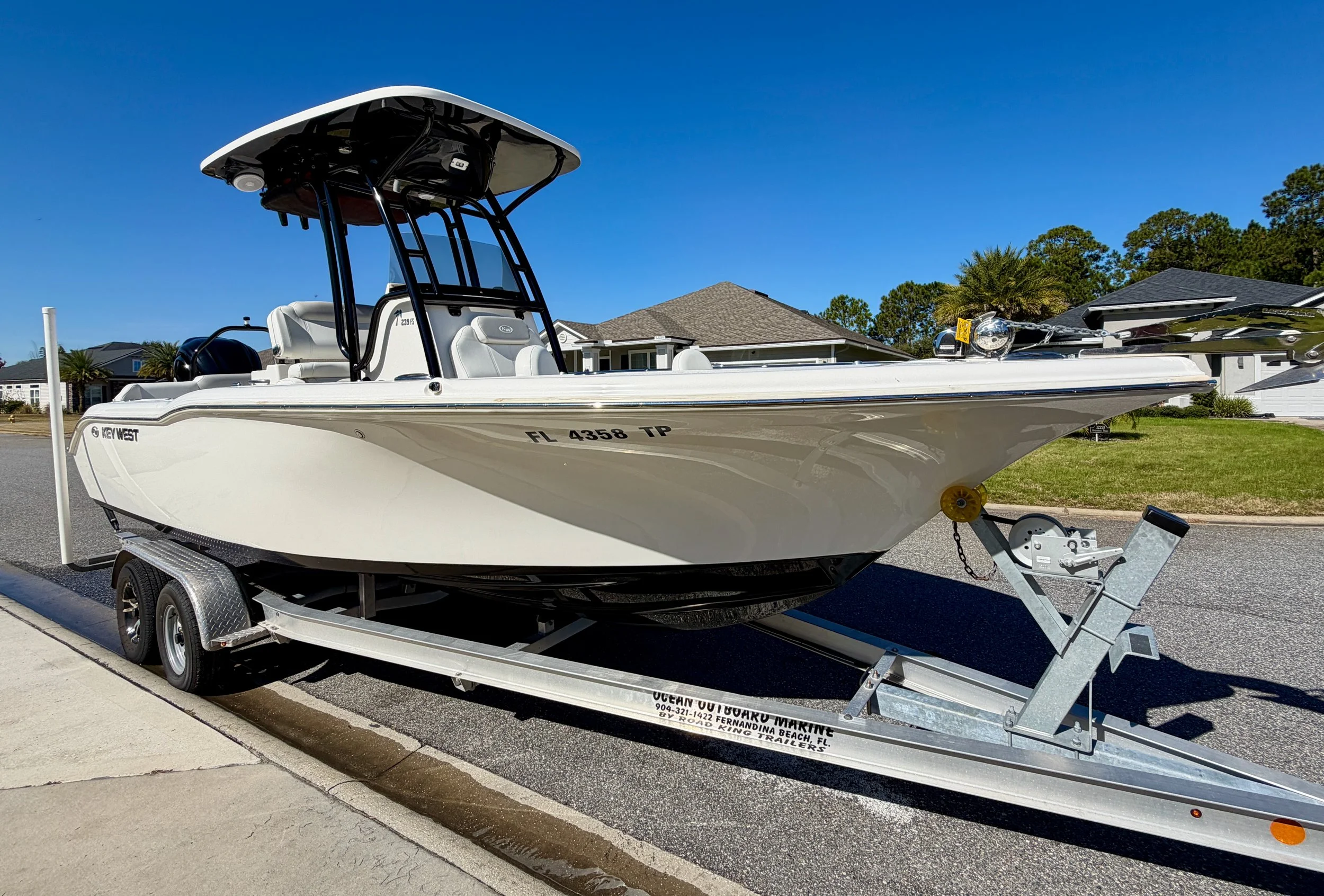 A white fishing boat on a trailer parked on a residential street with houses and trees in the background.