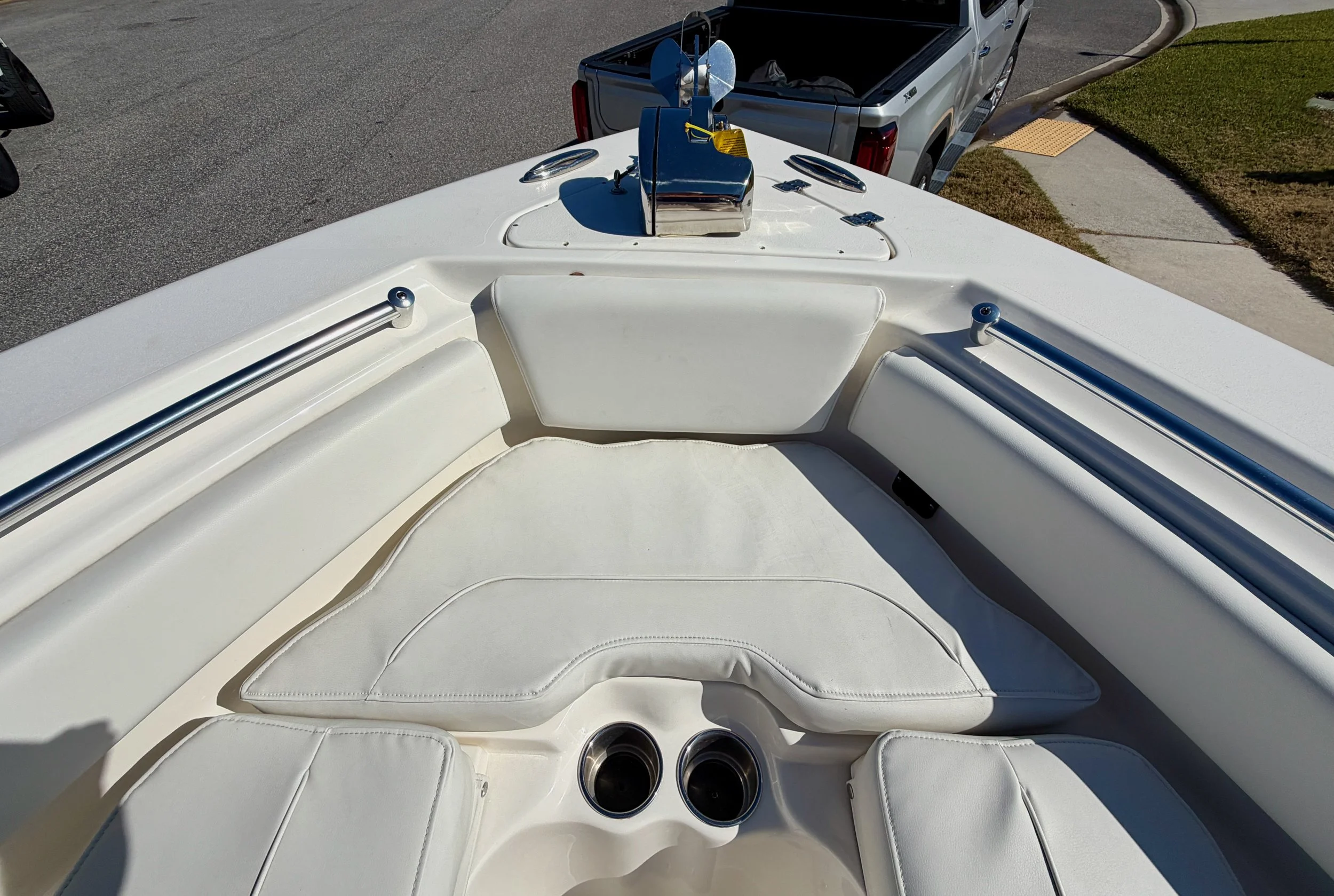 Front view of the bow of a white boat with cushioned seating and cup holders, parked on a street with a pickup truck in the background.