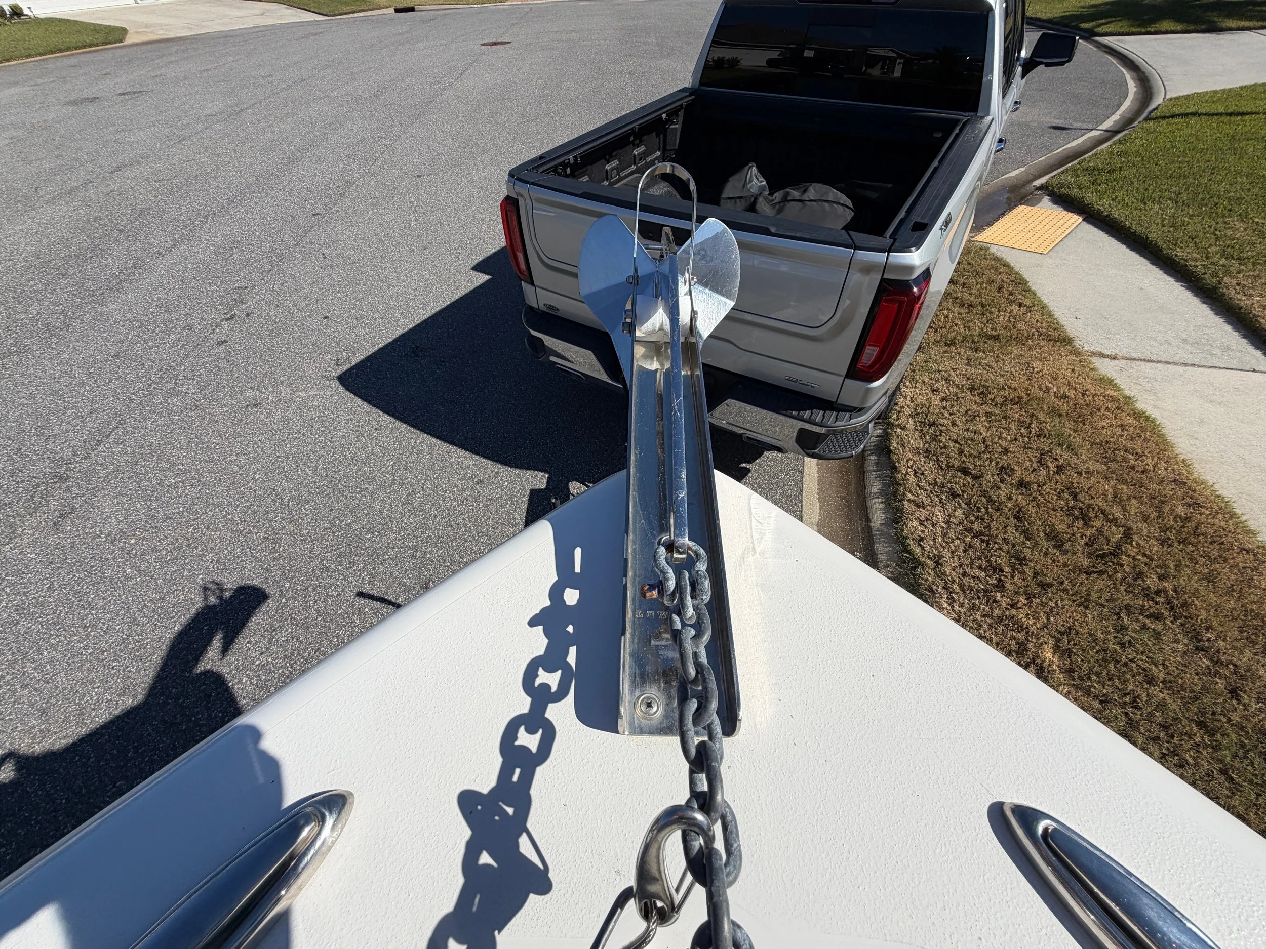 View from the front of a boat showing the anchor chain and anchor, with a pickup truck parked on the street beside the boat, and a sidewalk with greenery.