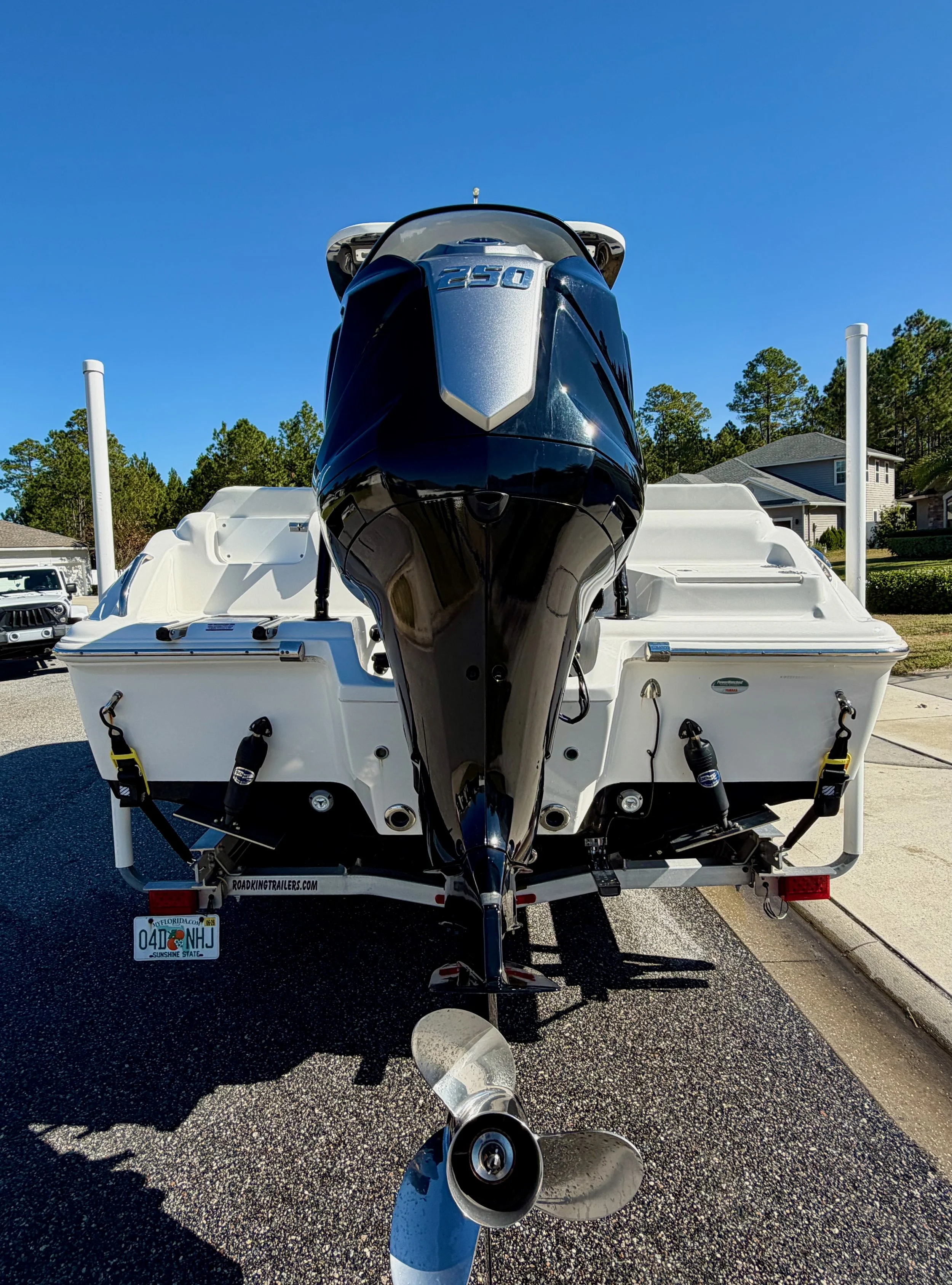 Front view of a white boat on a trailer, with a black and silver outboard motor labeled '250', parked on a paved area under a clear blue sky, in a residential neighborhood.
