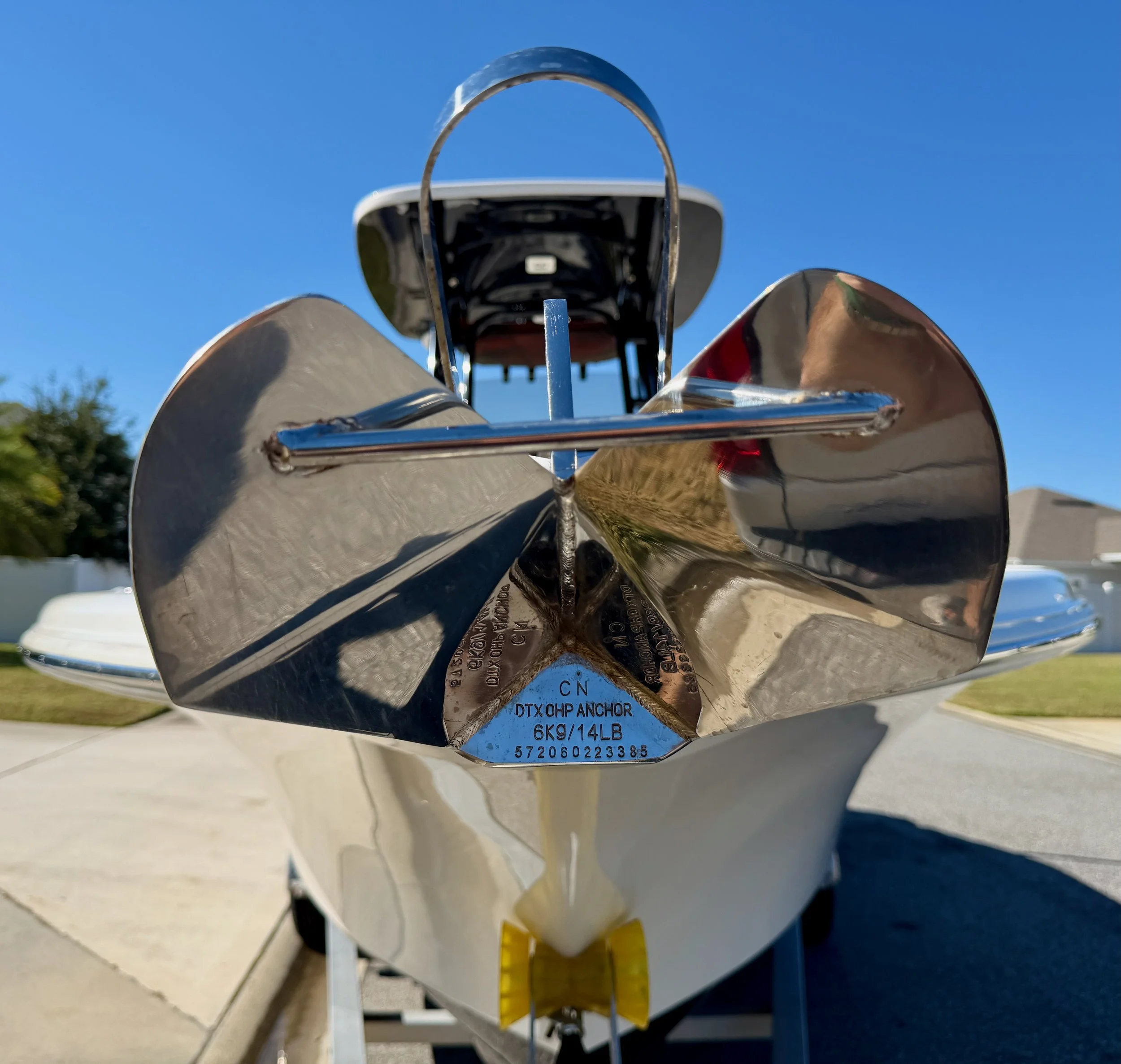 Close-up view of the bow of a small white boat with a shiny metal anchor attached, under a clear blue sky, with some houses and trees in the background.