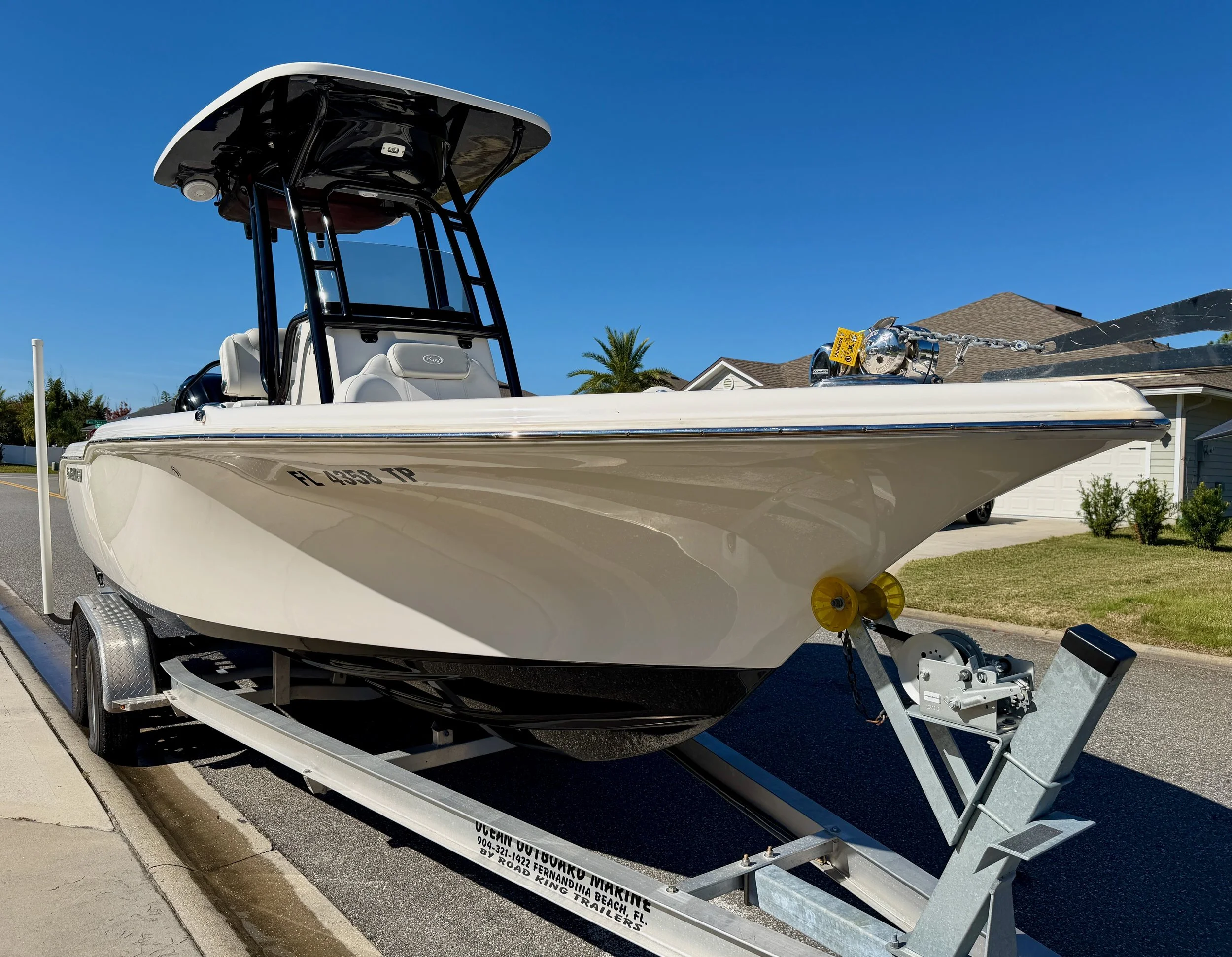A white center console fishing boat on a trailer parked on a suburban street under a blue sky.