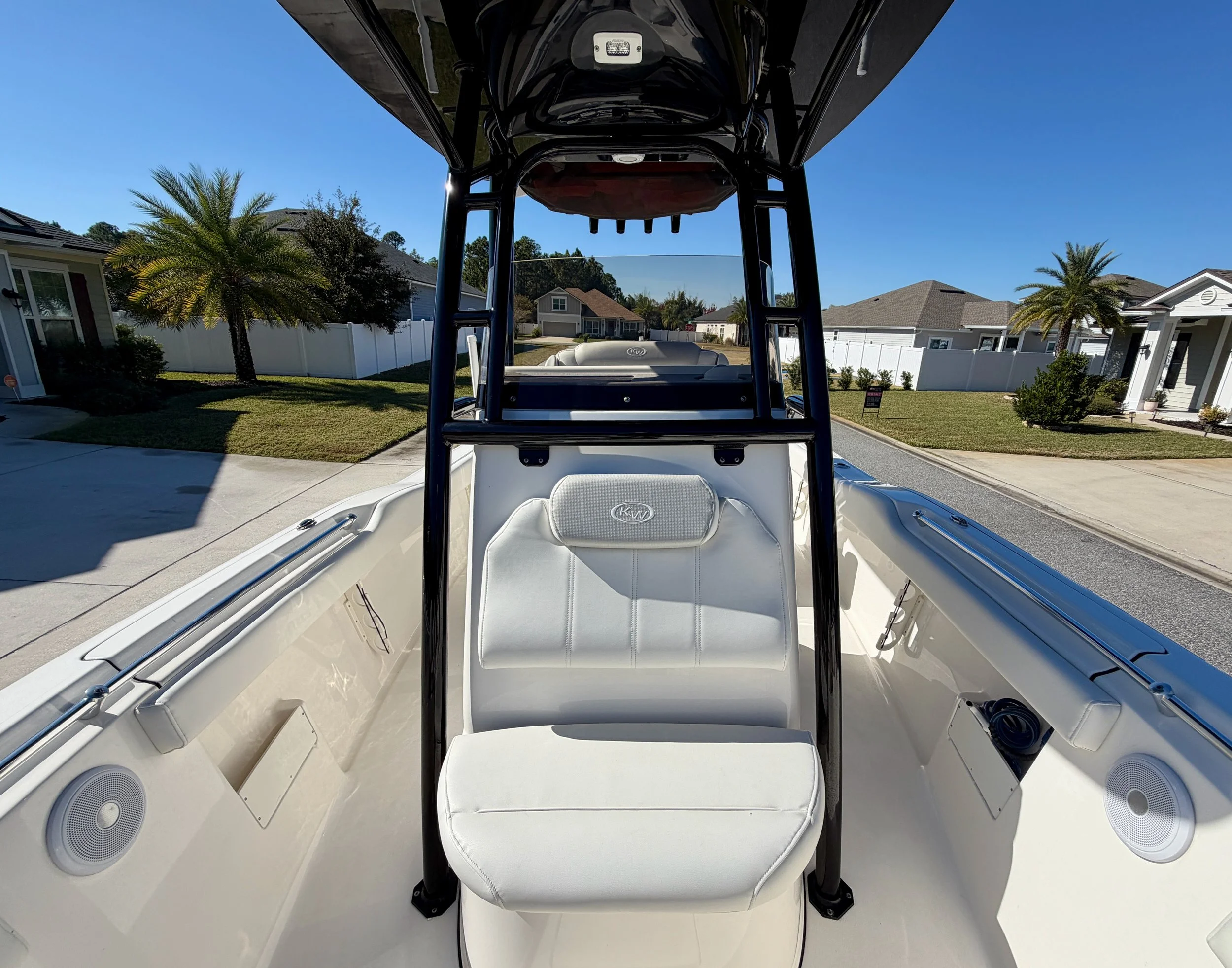 View from the front of a white boat with a black T-top, parked on a residential street with homes, trees, and a blue sky in the background.