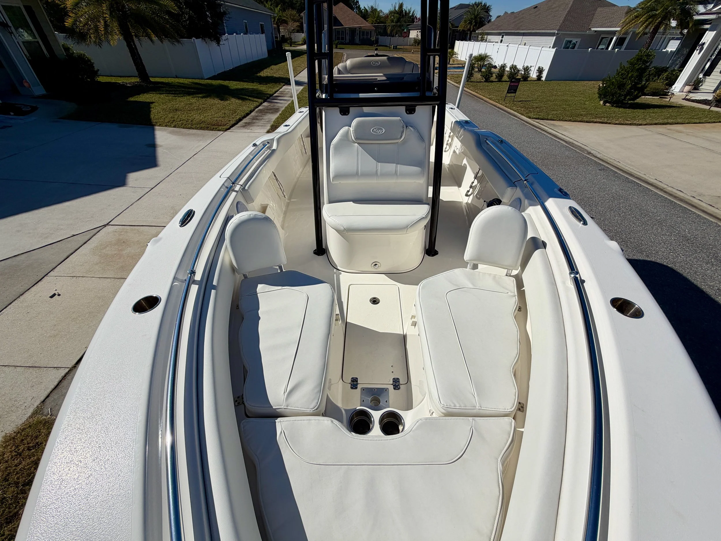 The interior of a white boat with cushioned seats and cup holders, parked on a driveway with houses and trees in the background.