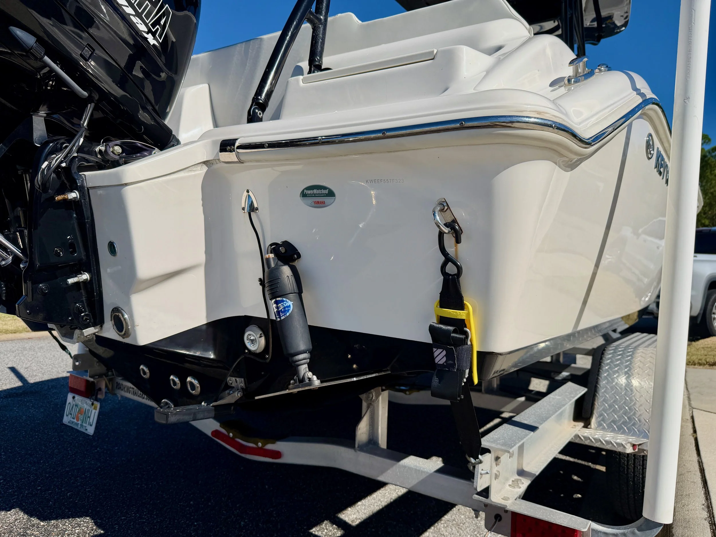 Close-up of the stern of a white motorboat on a trailer, showing outboard motor, tie-down straps, and a small sticker, with part of a vehicle and parking lot in the background.