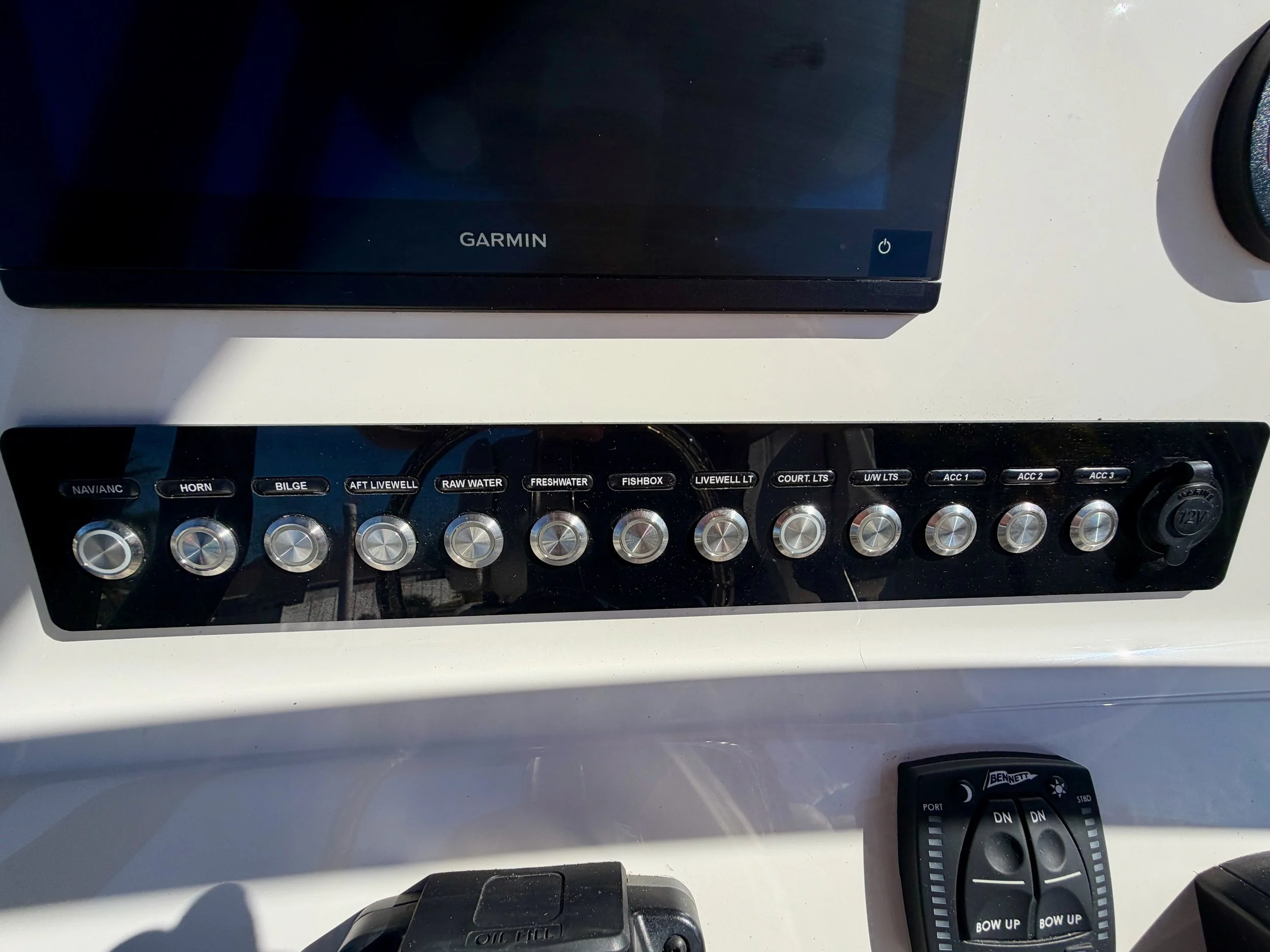 Boat control panel with various switches labeled for navigation, horn, bilge, livewell, raw water, freshwater, fishbox, livewell lights, court lights, underwater lights, and accessories, also showing a 12V power socket and another control device.